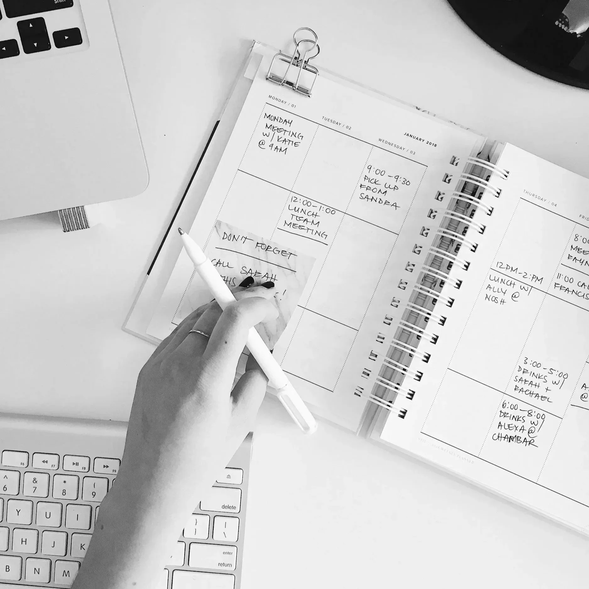 Black and white photo of a desk with a keyboard, a computer monitor, papers, a pen, and a person's hand holding a notepad with handwritten notes. The notes include scheduled meetings and reminders for the week. Specifically a midwife's calendar