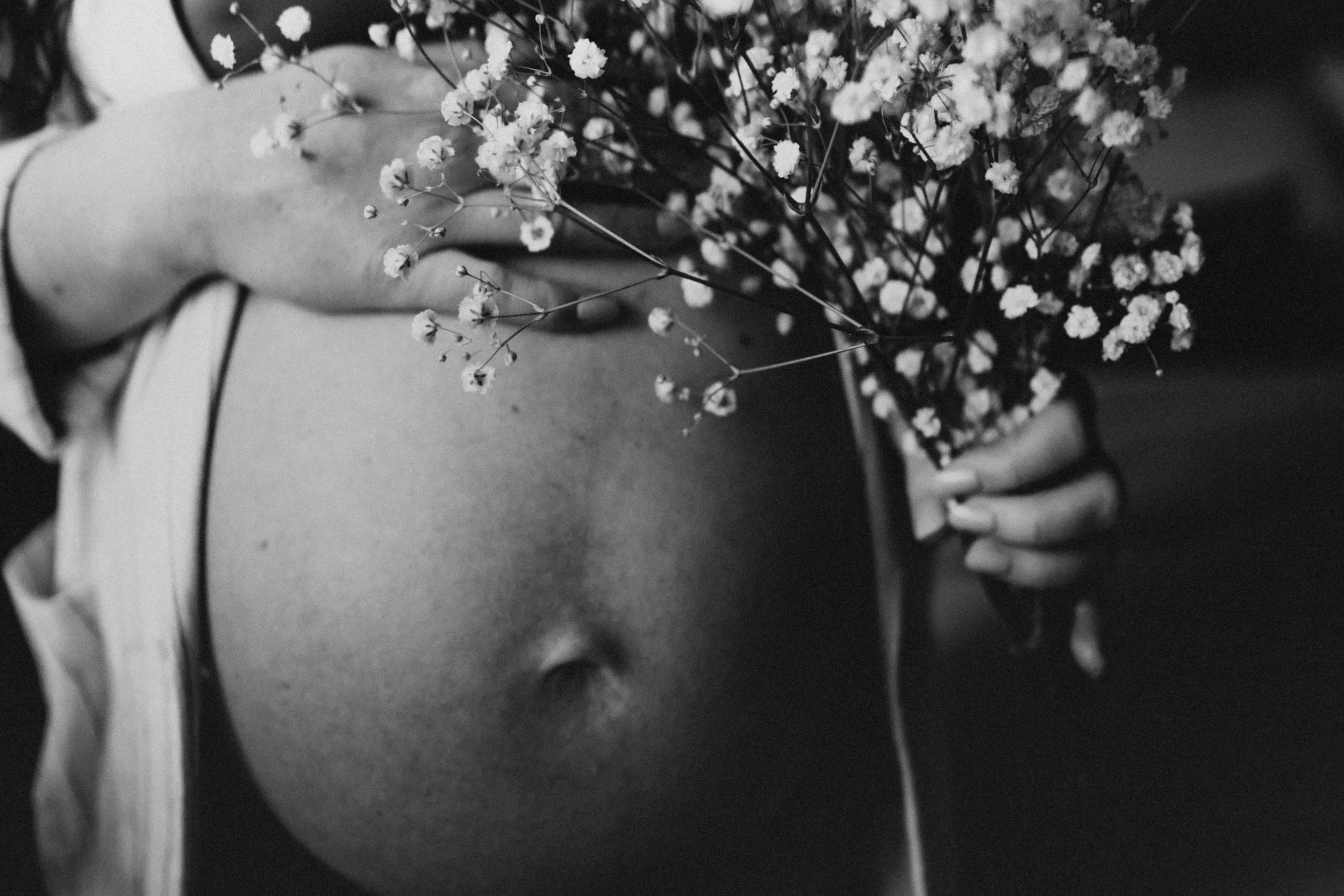 Black and white photo of a person's torso, with visible belly button and full pregnant belly, holding a bouquet of small flowers near the chest. The person has light skin and is wearing a strappy top.