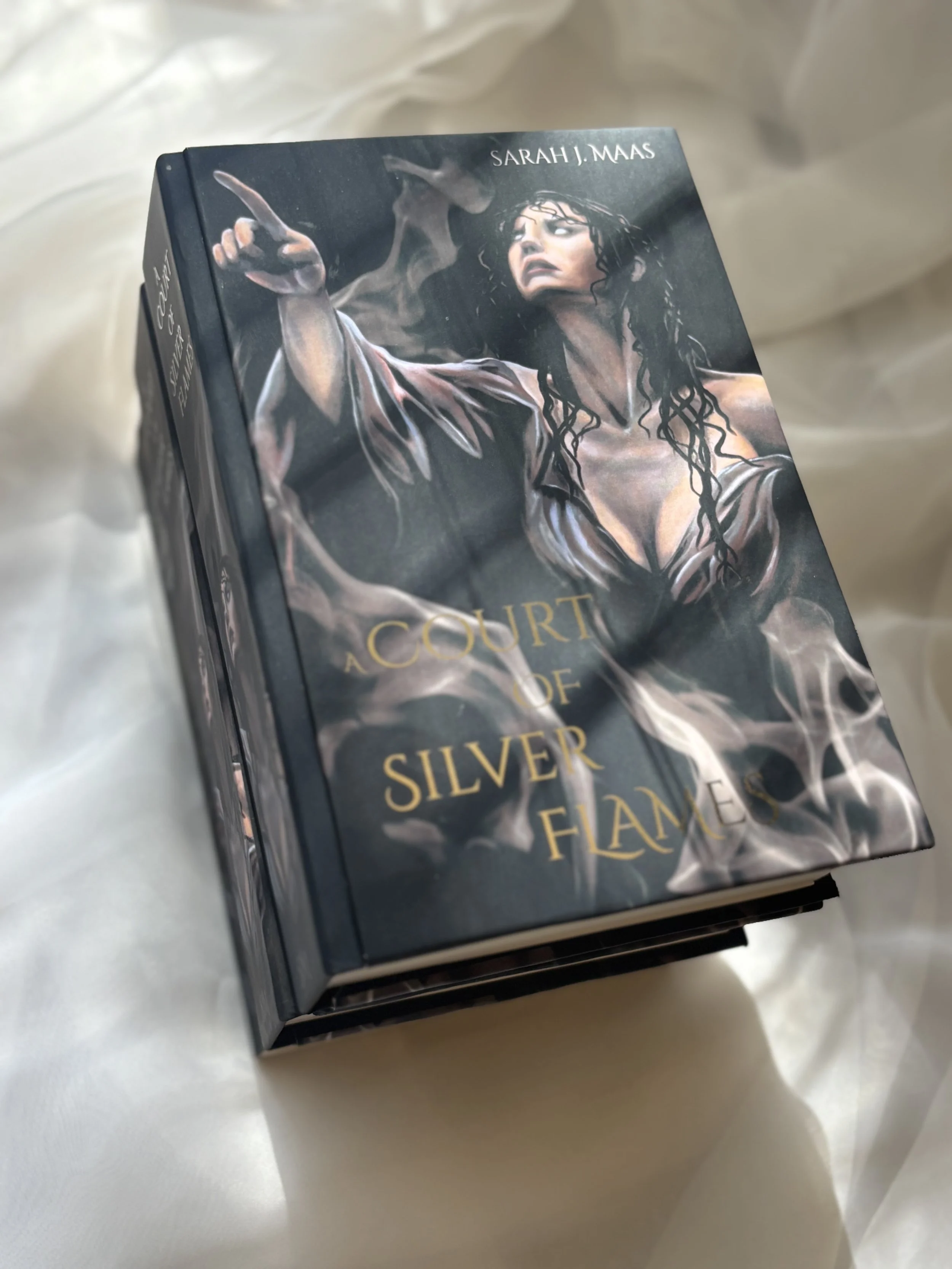 A special edition hardcover rebind (Firebound Books) of "A Court of Silver Flames" by Sarah J. Maas, featuring an exclusive art by Daphne Noel:  a woman with long dark hair, wearing a flowing dress, and pointing with her finger.