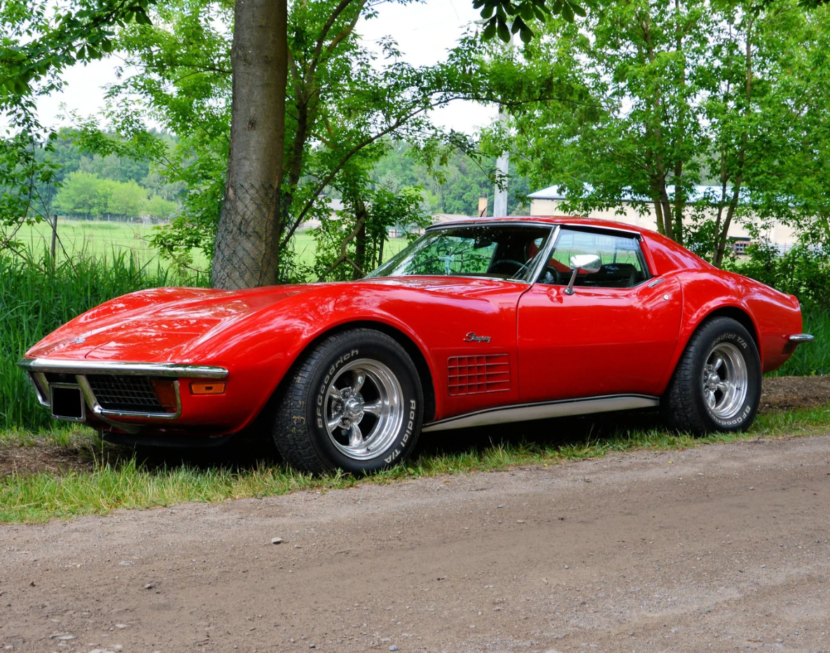 Red vintage sports car parked on a dirt road, surrounded by trees and greenery.