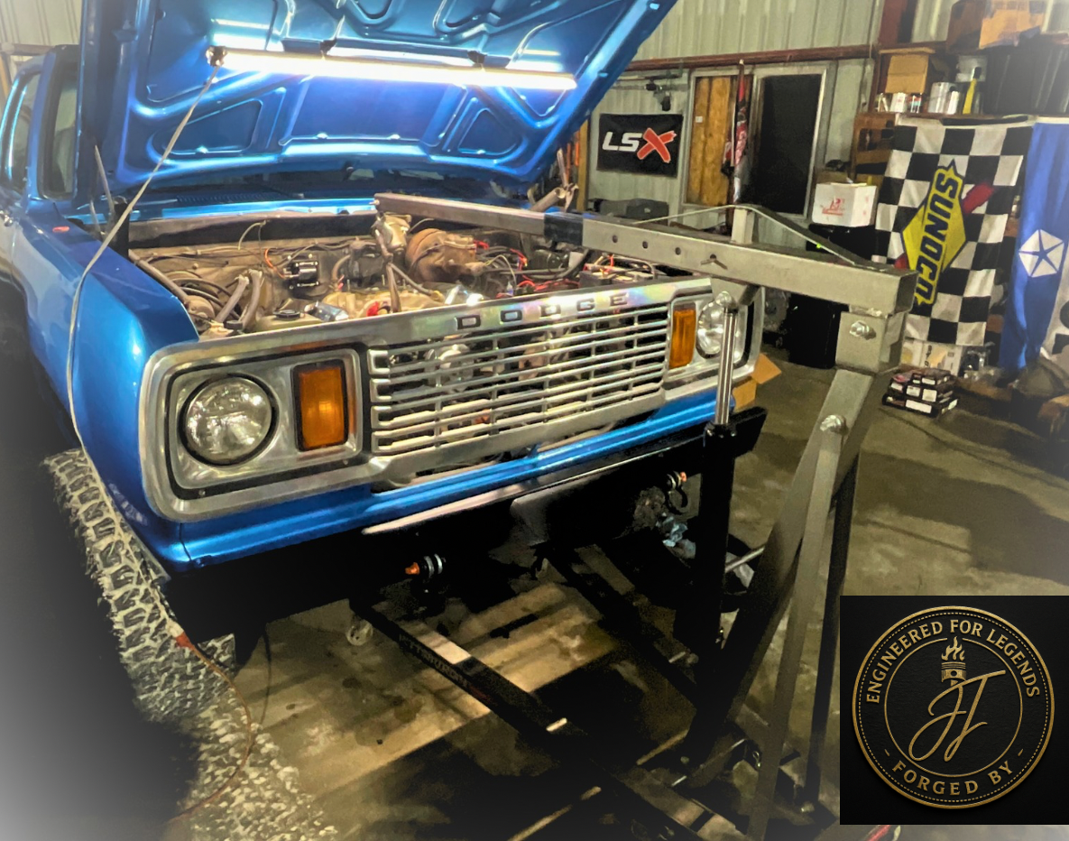 A blue classic Dodge pickup truck with the hood open in a garage setting, revealing the engine. Various automotive tools and banners are visible in the background. A circular logo with "Engineered for Legends, Forged by JF" is present in the image co