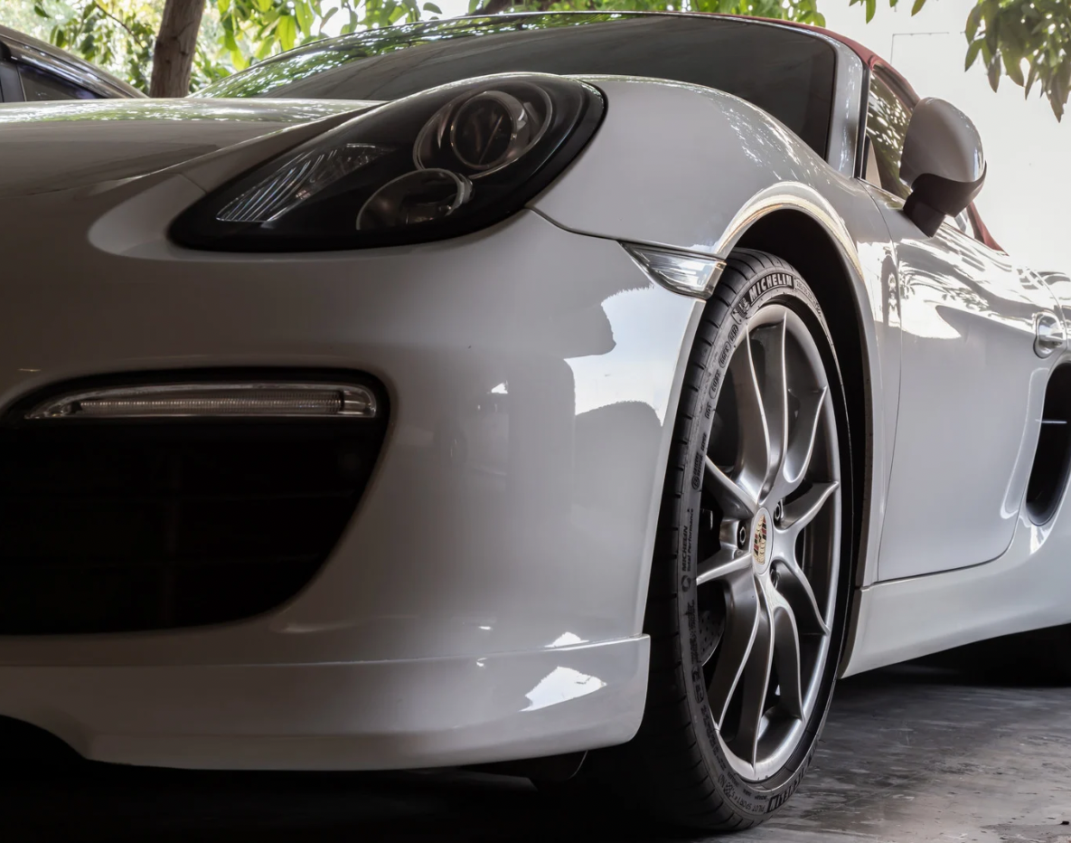 Close-up of a white sports car's front and side, featuring a sleek headlight, alloy wheel, and side mirror, parked under trees.