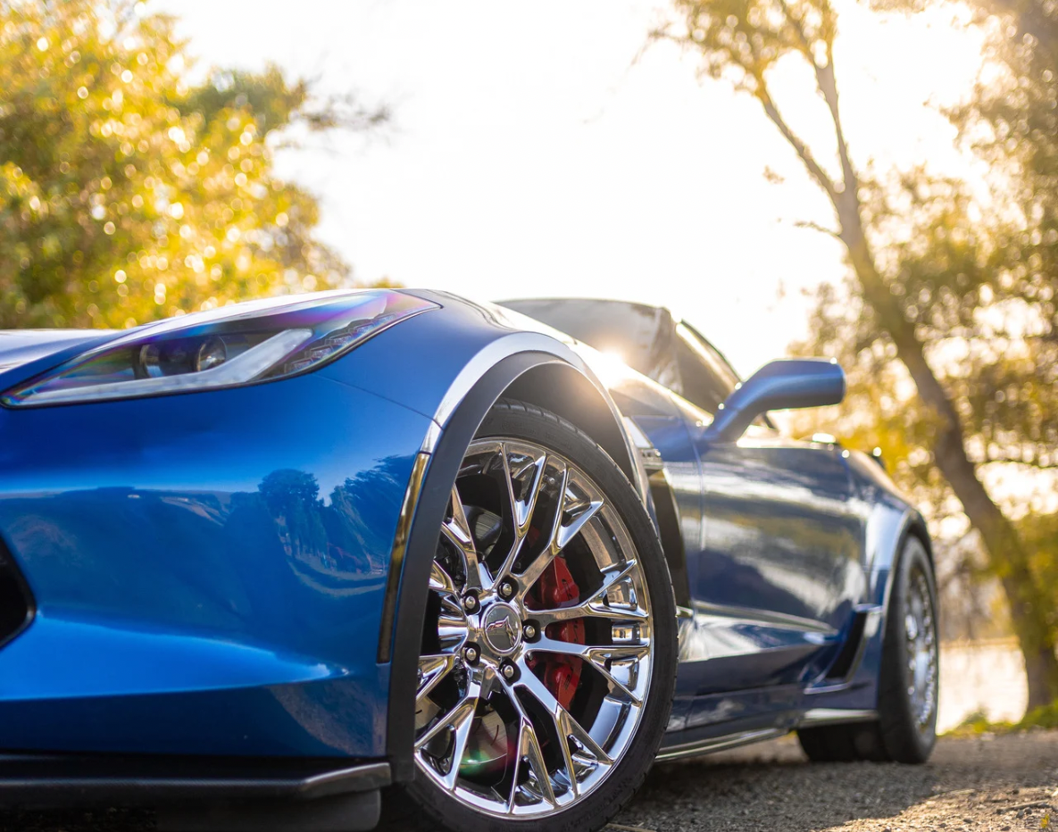 Close-up of a blue sports car with chrome wheels and a shining headlight, captured in a sunny outdoor setting with blurred trees in the background.