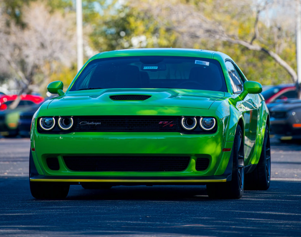 Front view of a vibrant green Dodge Challenger sports car parked on a street, surrounded by trees and other vehicles in the background.