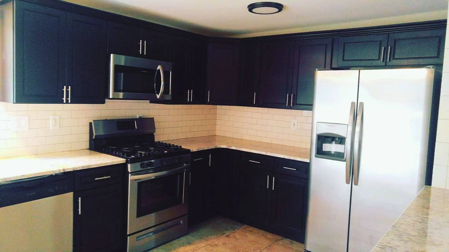 Kitchen featuring dark cabinets, a stainless steel microwave above a gas stove, a side-by-side stainless steel refrigerator, beige tile backsplash, and beige countertops.