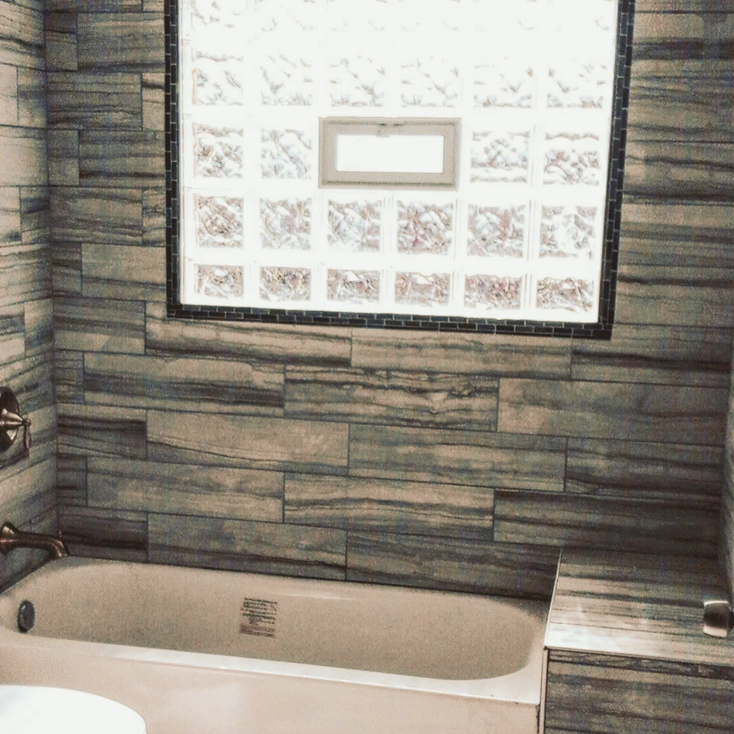 Bathroom with a bathtub, tiled gray wood-look walls, and a large glass block window.