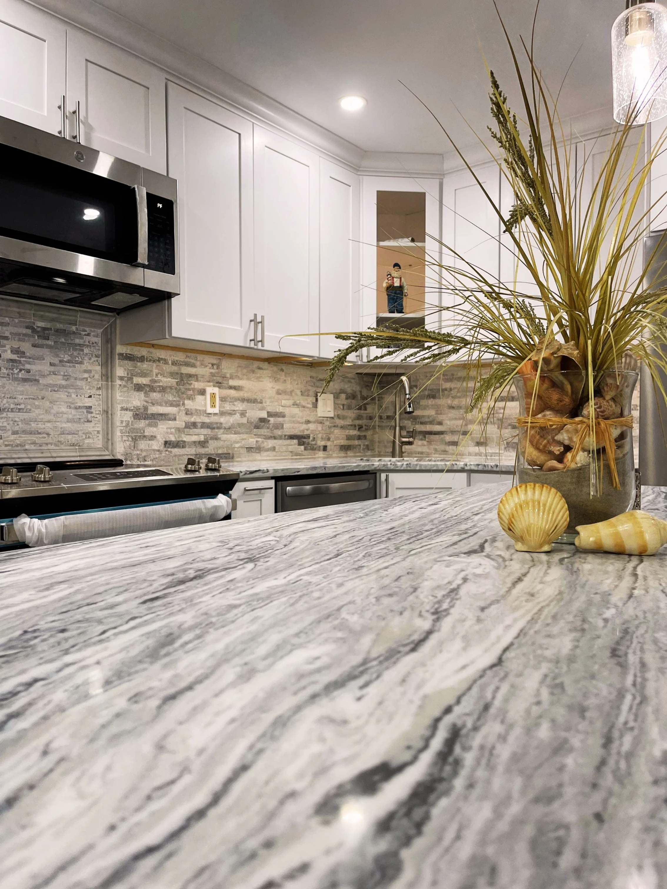 A modern kitchen with white cabinets, a gray stone backsplash, granite countertop, and a vase with dried plants and seashells on the counter.