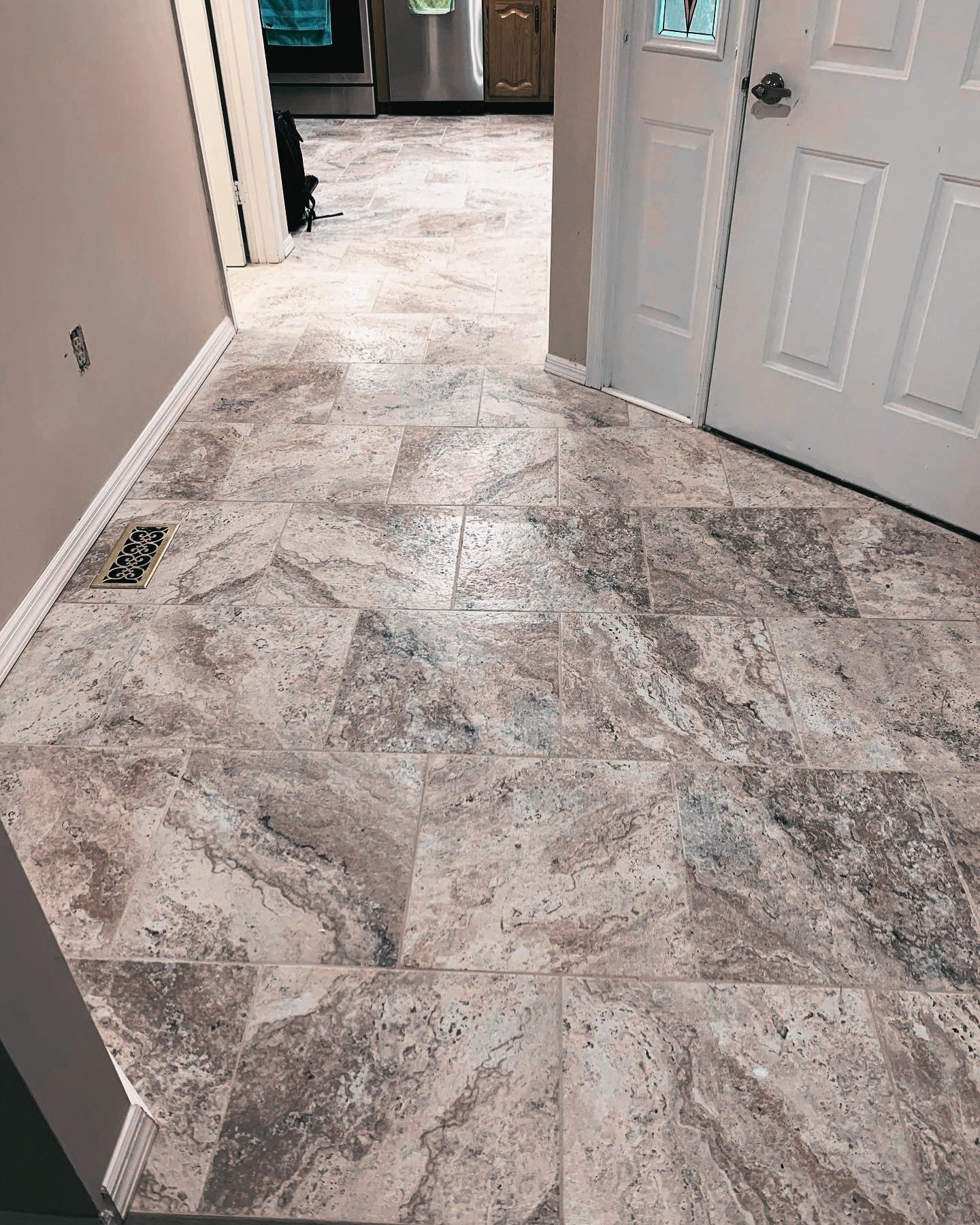 Interior view of a tiled floor in a home, with a door on the right and an opening leading to a kitchen area with cabinets and appliances in the background.