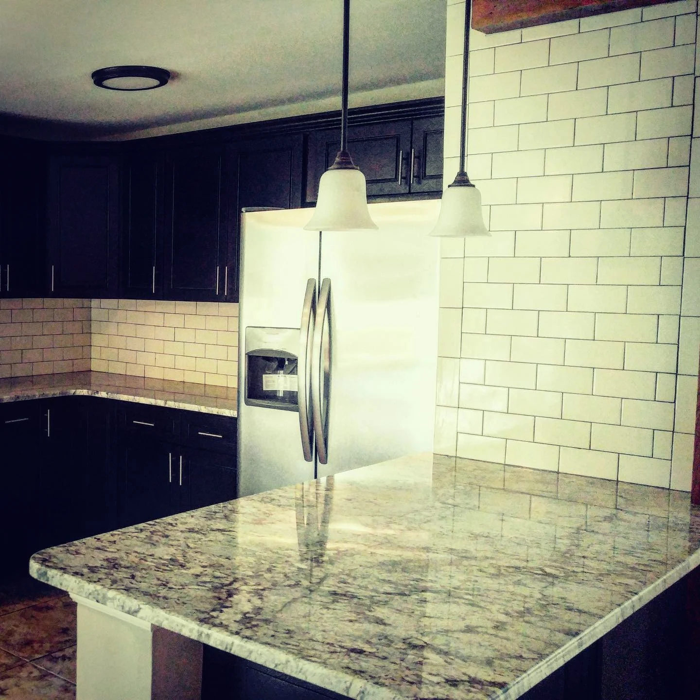 Kitchen with dark wooden cabinets, a granite countertop, a stainless steel refrigerator, and hanging pendant lights over the counter.