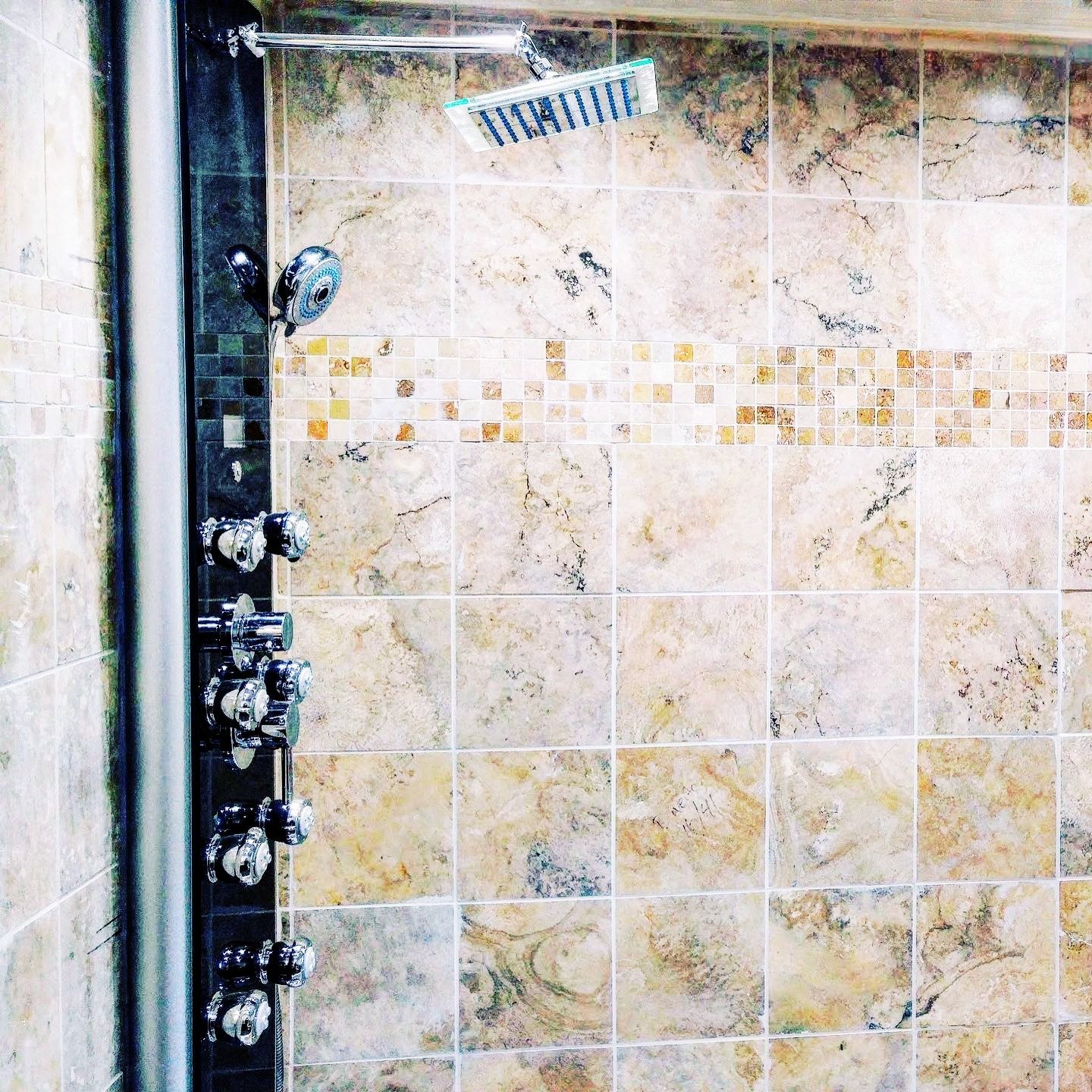 Shower with multiple chrome knobs, a black shower panel, a handheld shower head, and a rain shower head on the ceiling. The wall has large beige marble tiles with a strip of small mosaic tiles.