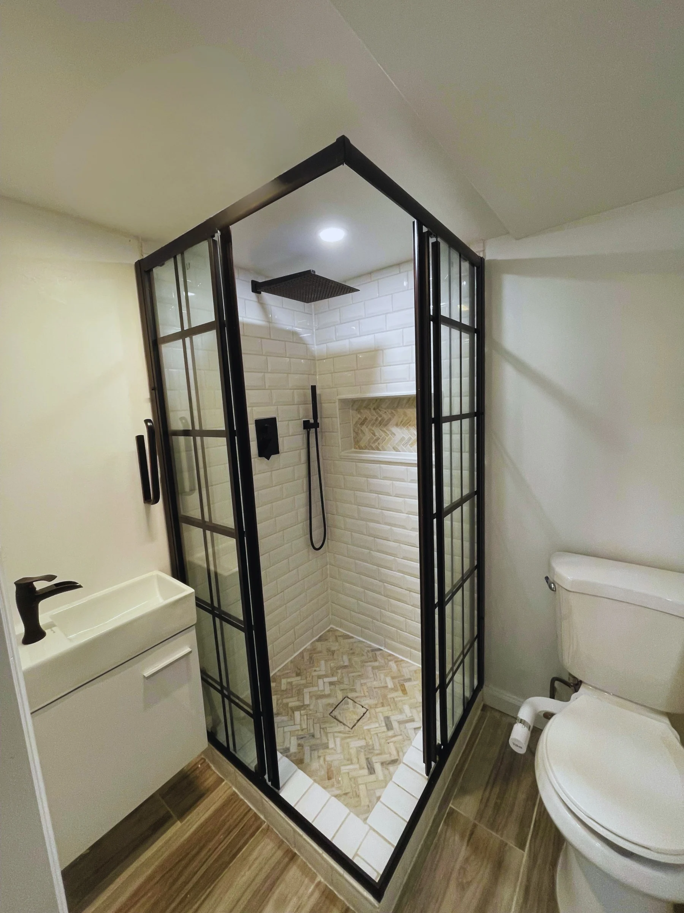 Small bathroom with a glass-enclosed shower, a toilet, and a small white sink with a black faucet. The shower has a black frame and a black showerhead, with white subway tiles on the walls and a herringbone pattern on the floor.