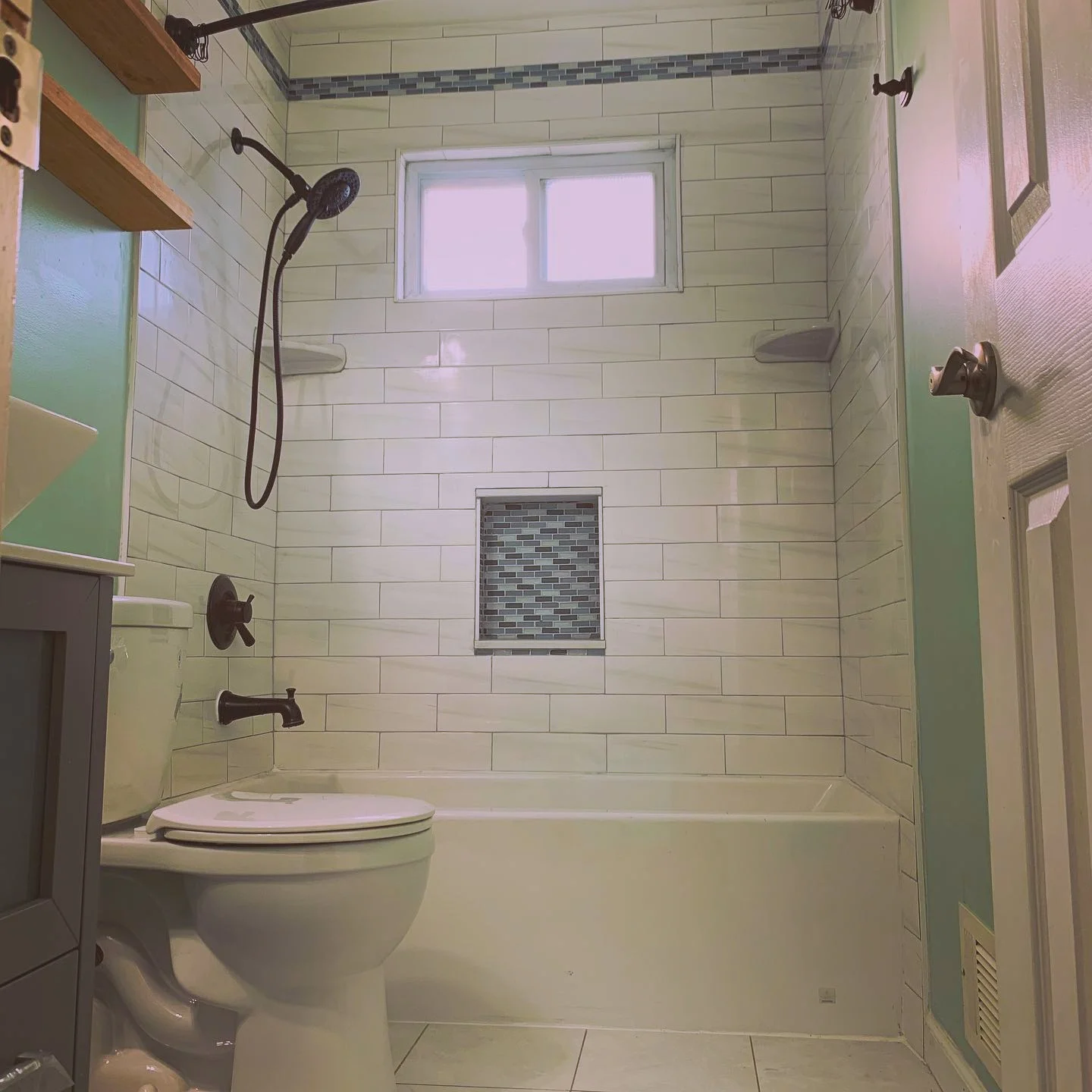 A bathroom with a bathtub, a shower with a handheld showerhead, a window, a toilet, and tiled walls with a decorative strip near the ceiling.