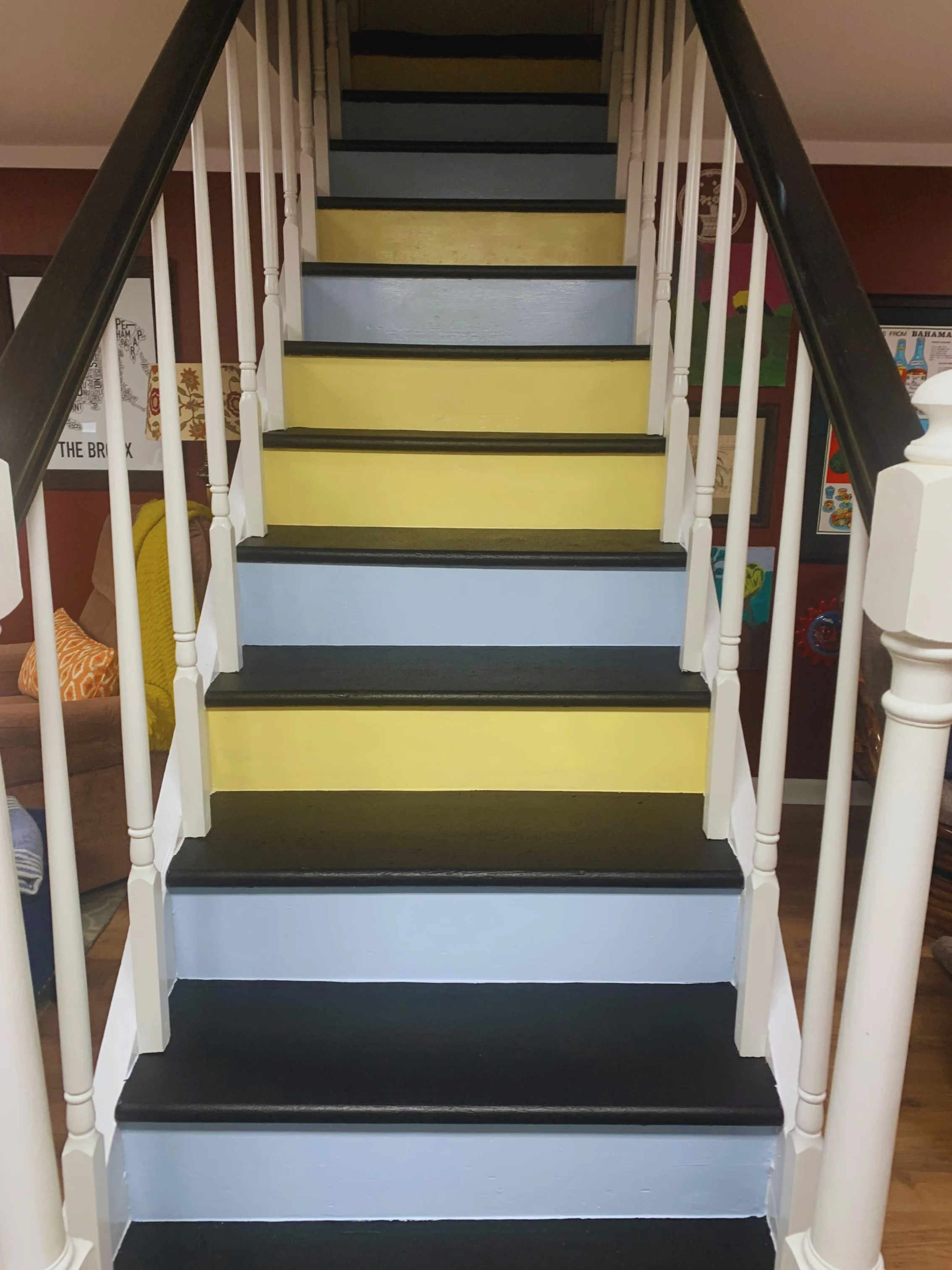 Colorful staircase with black treads, yellow risers, and white balustrades, leading upstairs in a room decorated with framed artwork on the walls.