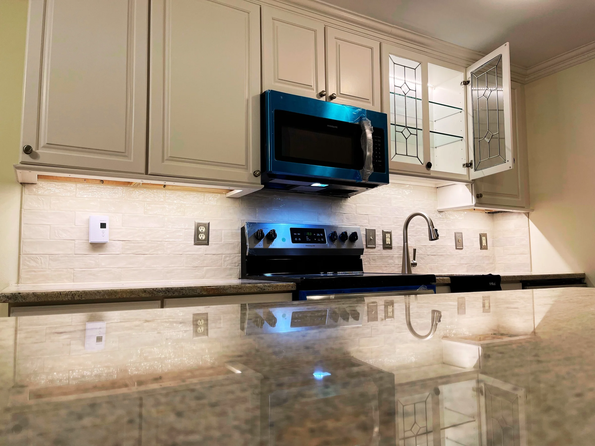 View of a kitchen with beige cabinets, a granite countertop, a black stove, a microwave oven with blue wrapping, a glass-front cabinet, and a stainless steel sink with a curved faucet. There are electrical outlets and switches on the backsplash.