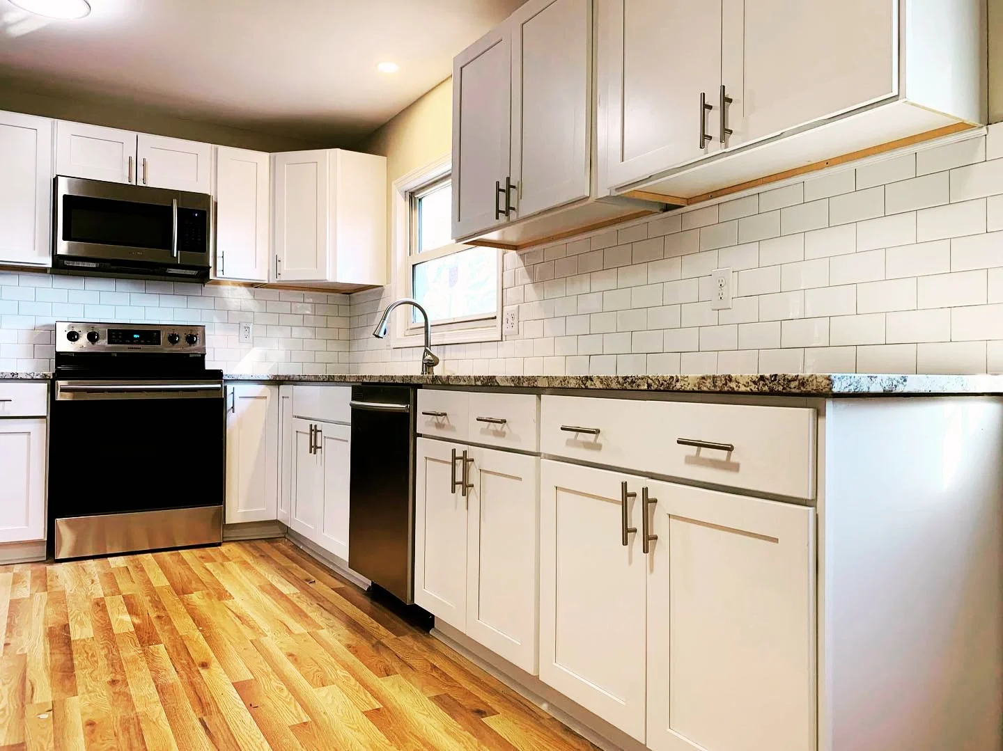 Kitchen with white cabinets, granite countertops, a white subway tile backsplash, a microwave above a black oven, a window above the sink, and wood flooring.