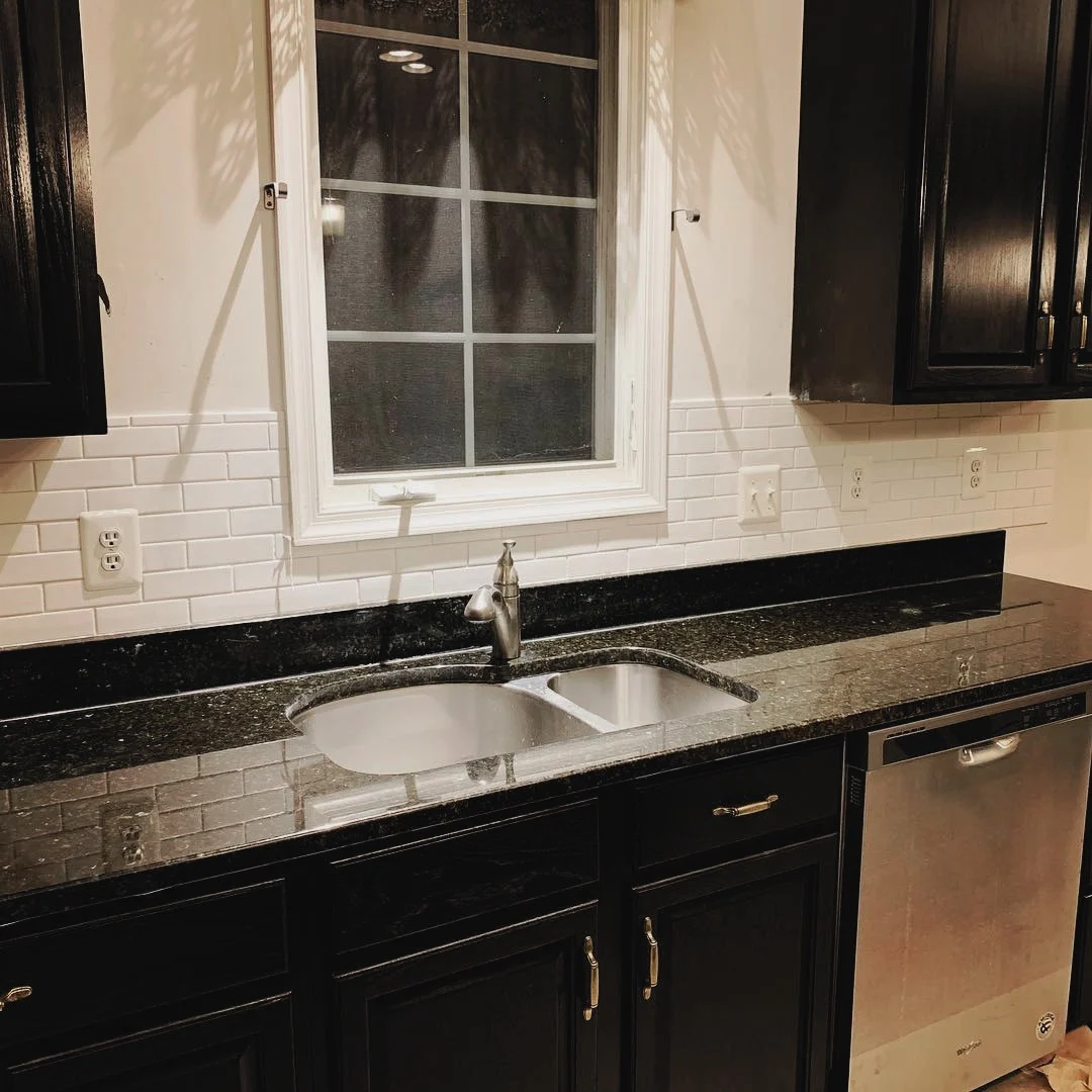 Kitchen sink area with black cabinets and granite countertop, a window with white trim, white subway tile backsplash, and a stainless steel dishwasher.