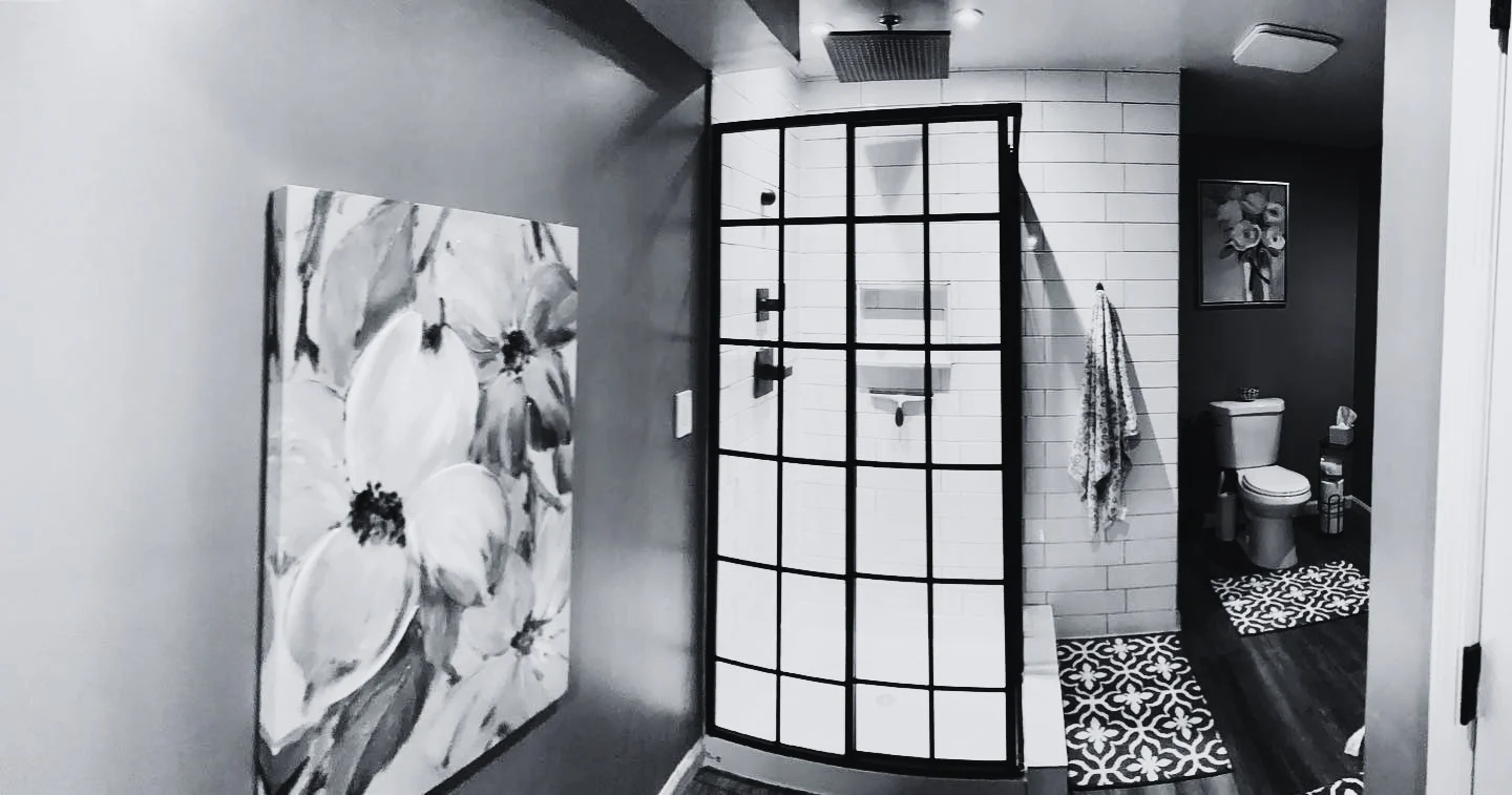 Bathroom with a glass shower enclosure, wall-mounted shower head, a toilet, and decorative rugs. Wall decor includes floral paintings and towels hanging on hooks.