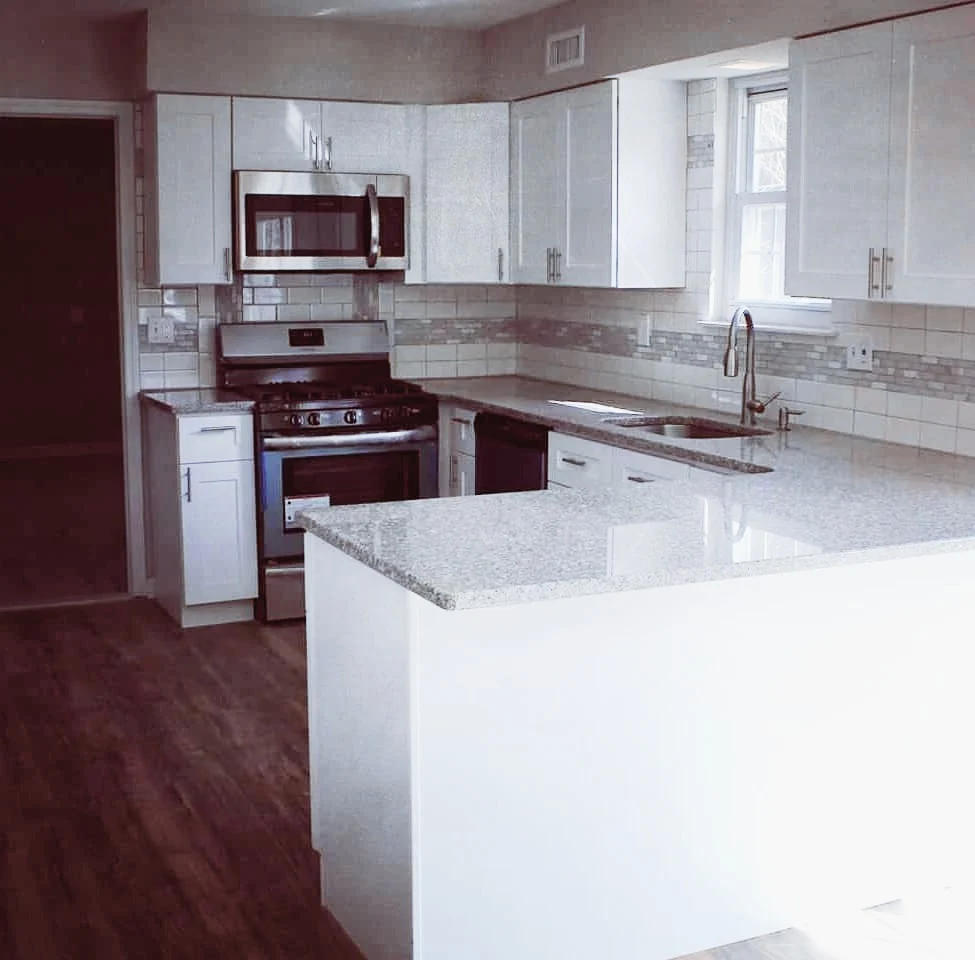 Modern kitchen with white cabinets, stainless steel microwave and oven, granite countertops, a window above the sink, and wooden flooring.