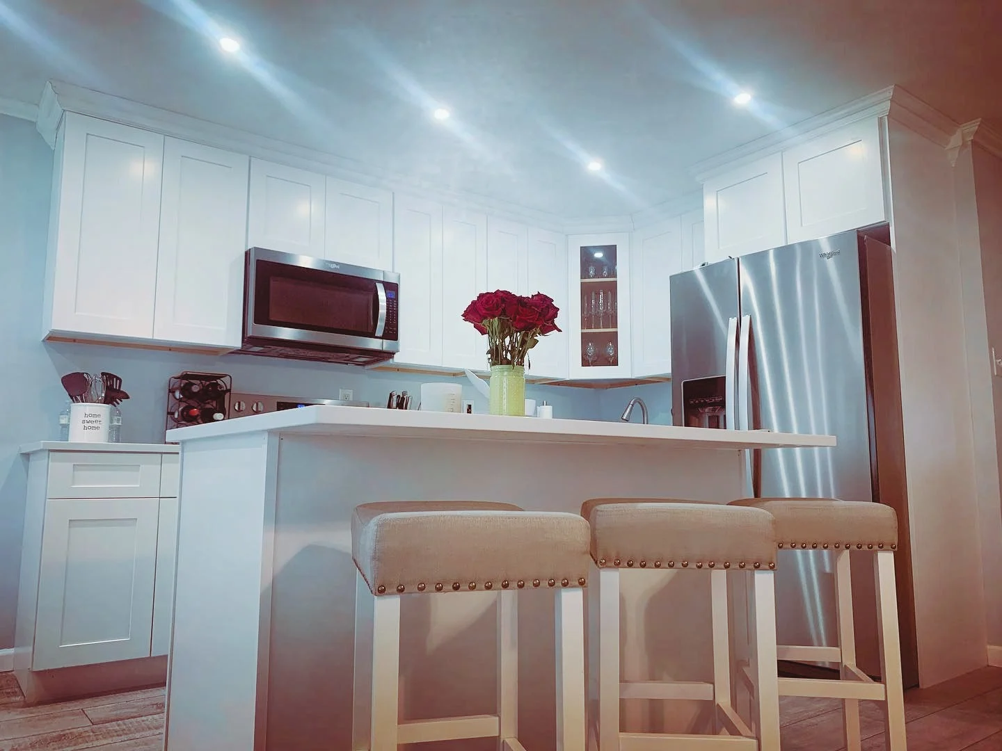 Modern kitchen with white cabinets, stainless steel refrigerator, microwave, and a kitchen island with a vase of red roses and three beige barstools.