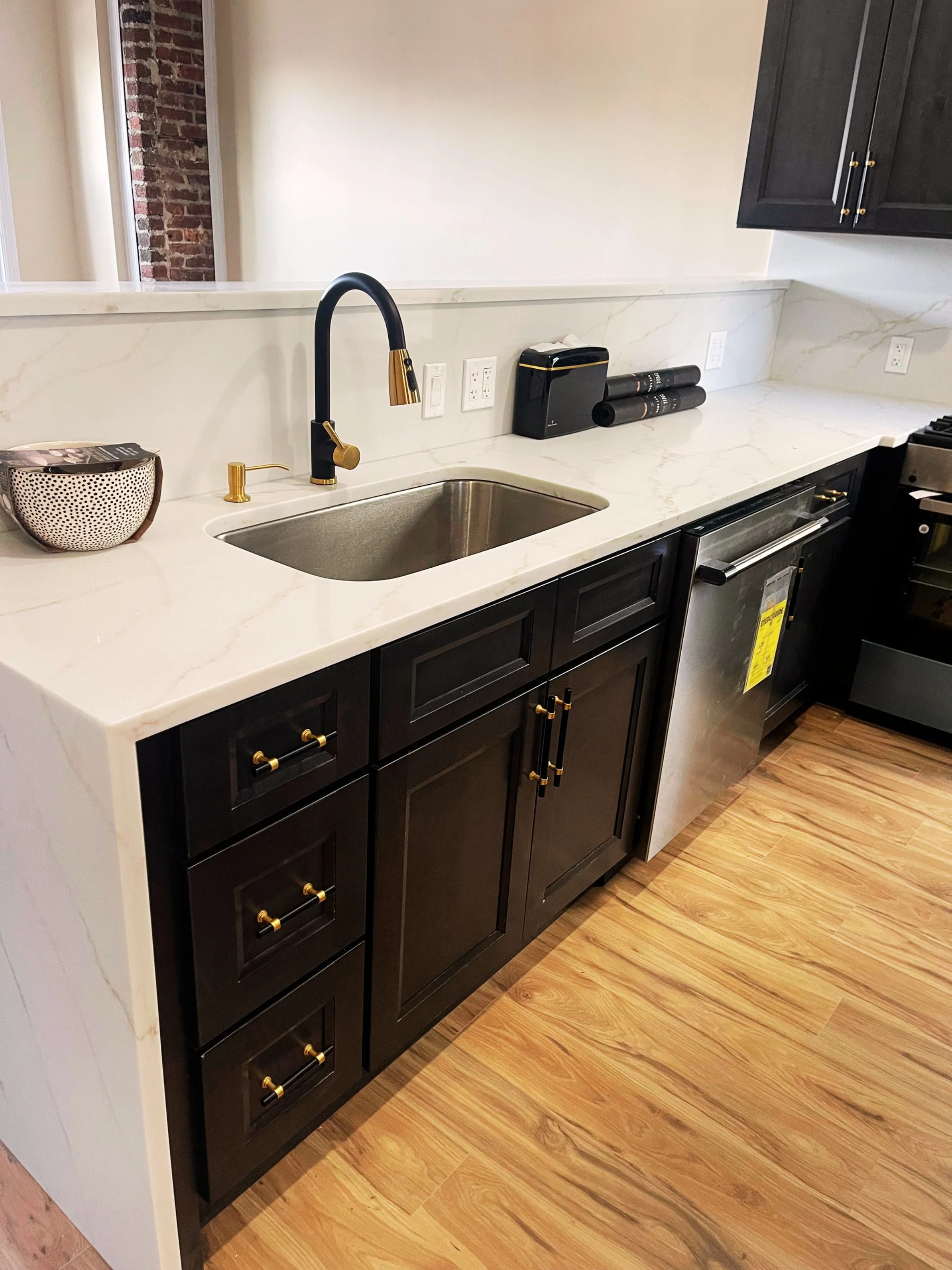 Modern kitchen with white marble countertop, black cabinets with gold handles, a stainless steel sink, black and gold faucet, and wooden flooring.