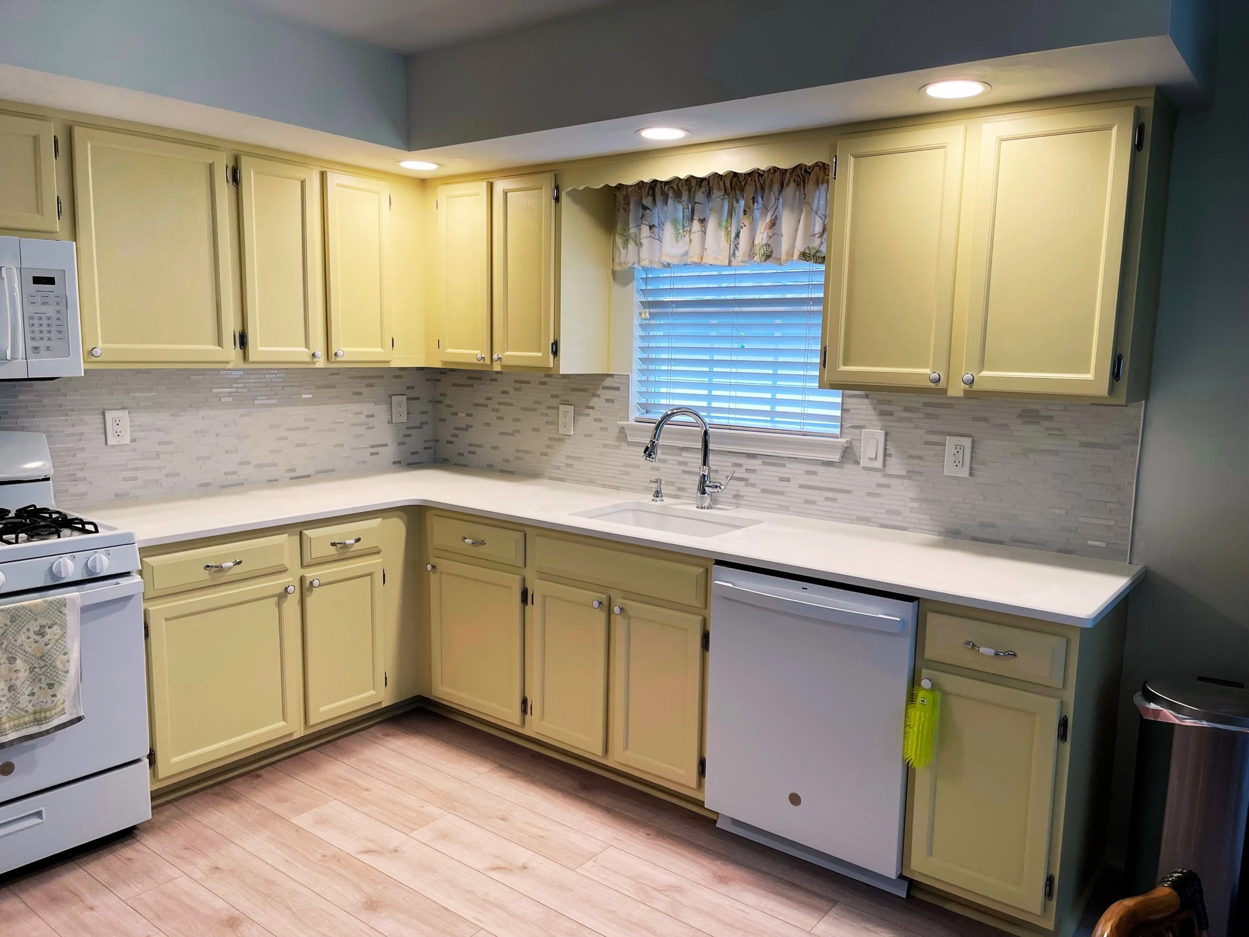 Bright yellow kitchen cabinets with white countertops, a white stove, a microwave, a window with blinds and a floral valance, a stainless steel sink with a faucet, a white dishwasher, and a stainless steel trash can in the corner.
