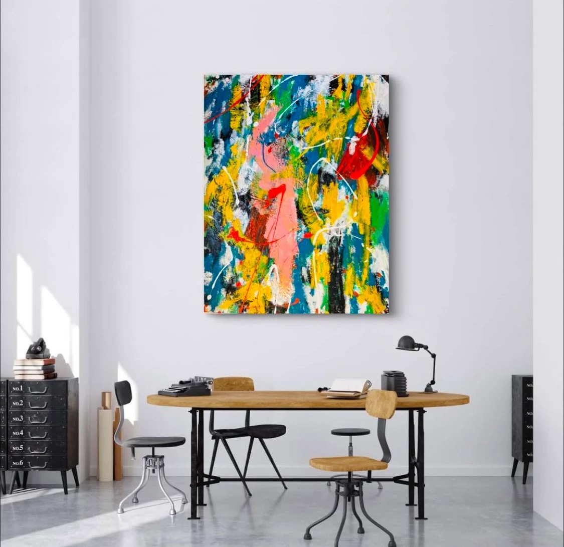 An abstract painting with vibrant colors hanging on a plain white wall in an office space. The room has a wooden desk, two chairs, and black storage units.