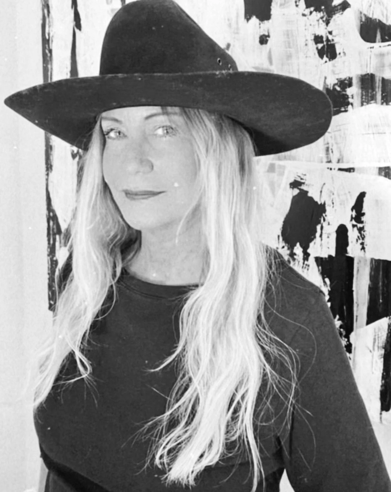 Black and white photo of a woman with long hair wearing a wide-brimmed hat and dark shirt, standing in front of an abstract painted background.