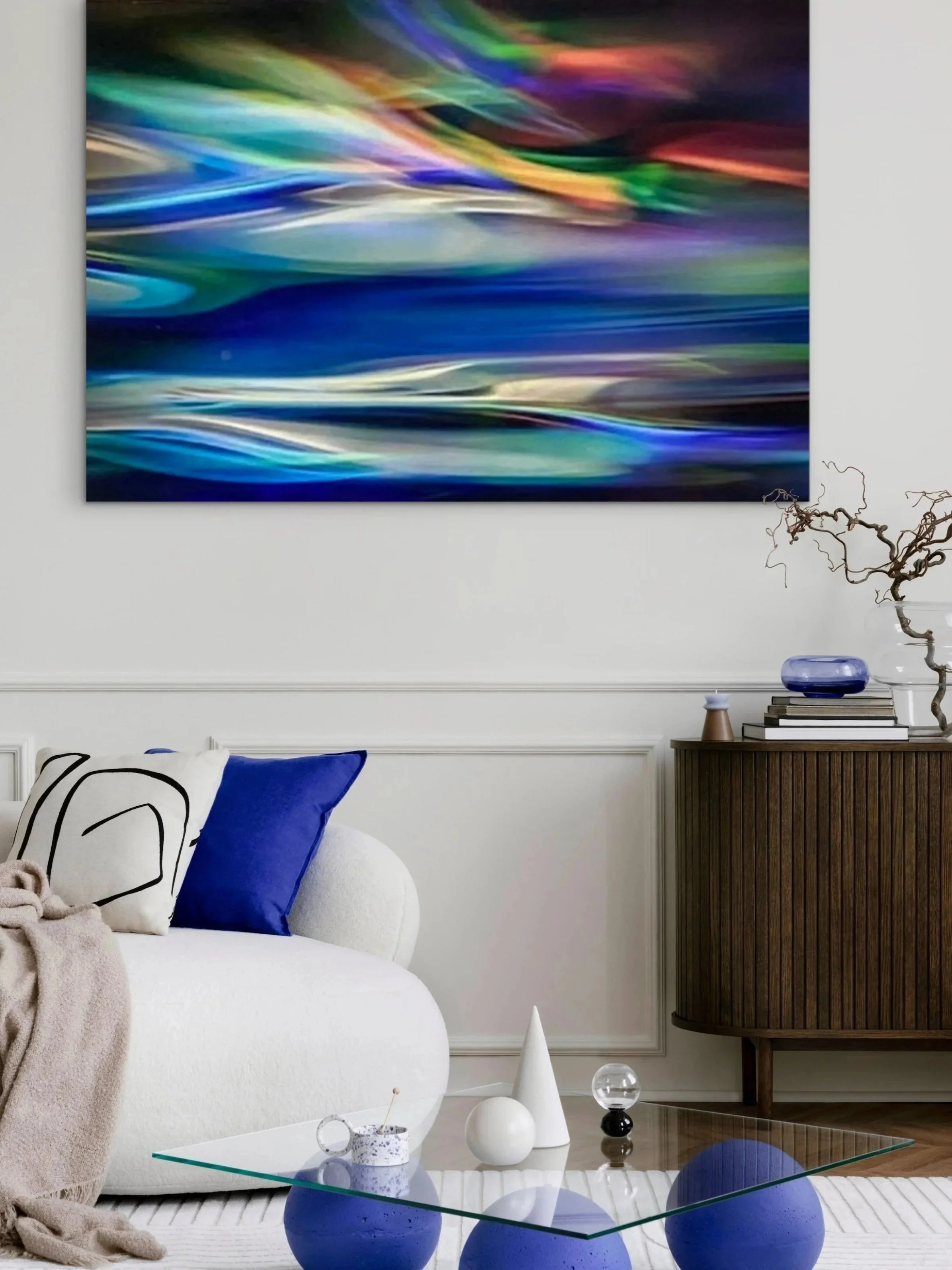 Living room with abstract colorful artwork on the wall, white sofa with navy and decorated pillows, glass coffee table with blue, white, and clear decorative objects, and a dark wooden sideboard with a glass bowl, a stack of books, and decorative branches.