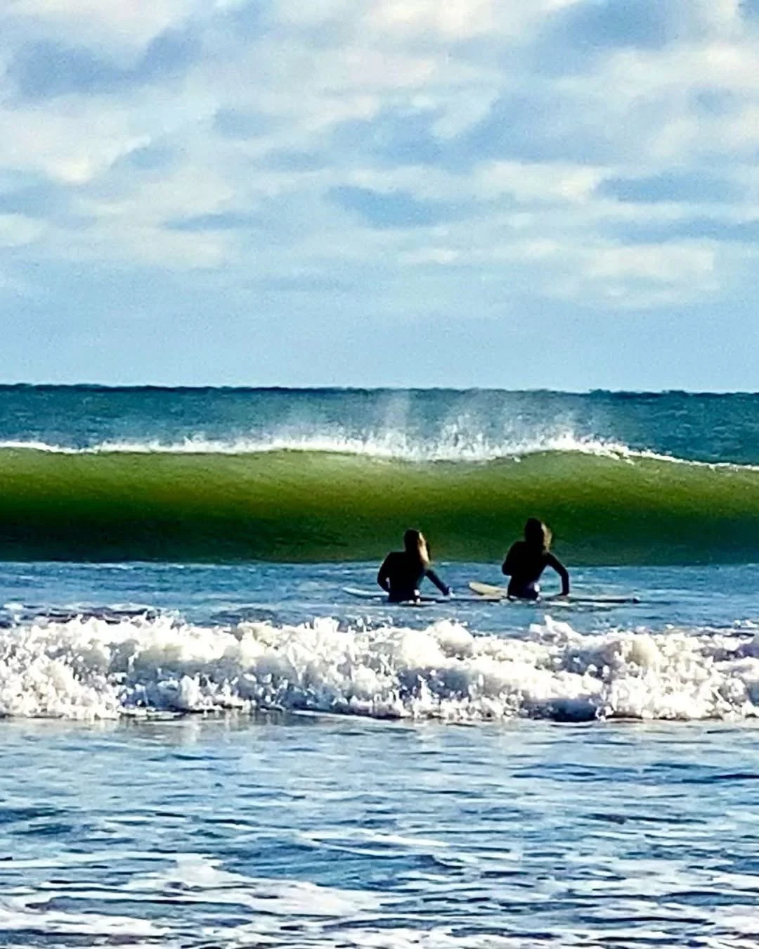 Two surfers sitting on their surfboards in the ocean, facing a large green wave in New Smyrna Beach, FL