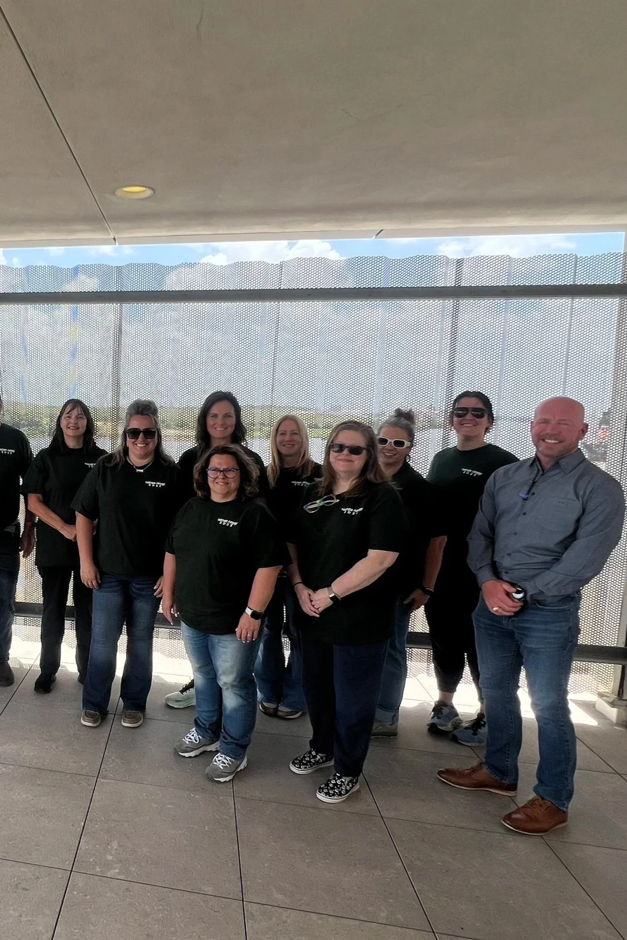 Group of smiling people, eight women and one man, standing on a balcony with a perforated metal wall behind them. The women are wearing black shirts, and the man is wearing a gray button-up shirt. The background shows a partly cloudy sky and distant landscape.