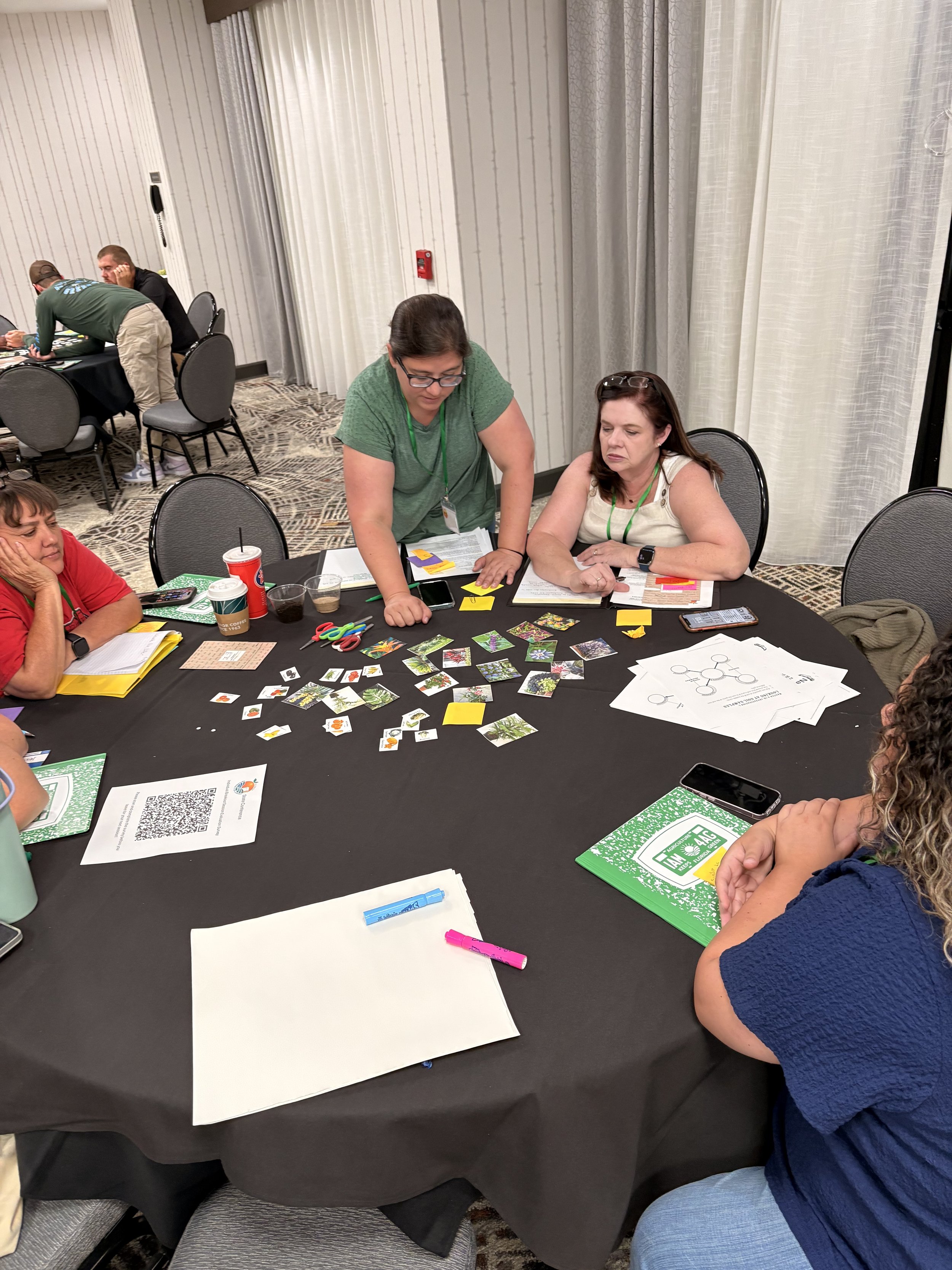 Teachers working around a black round table with office supplies, photos, papers, and snacks during a meeting or team activity.