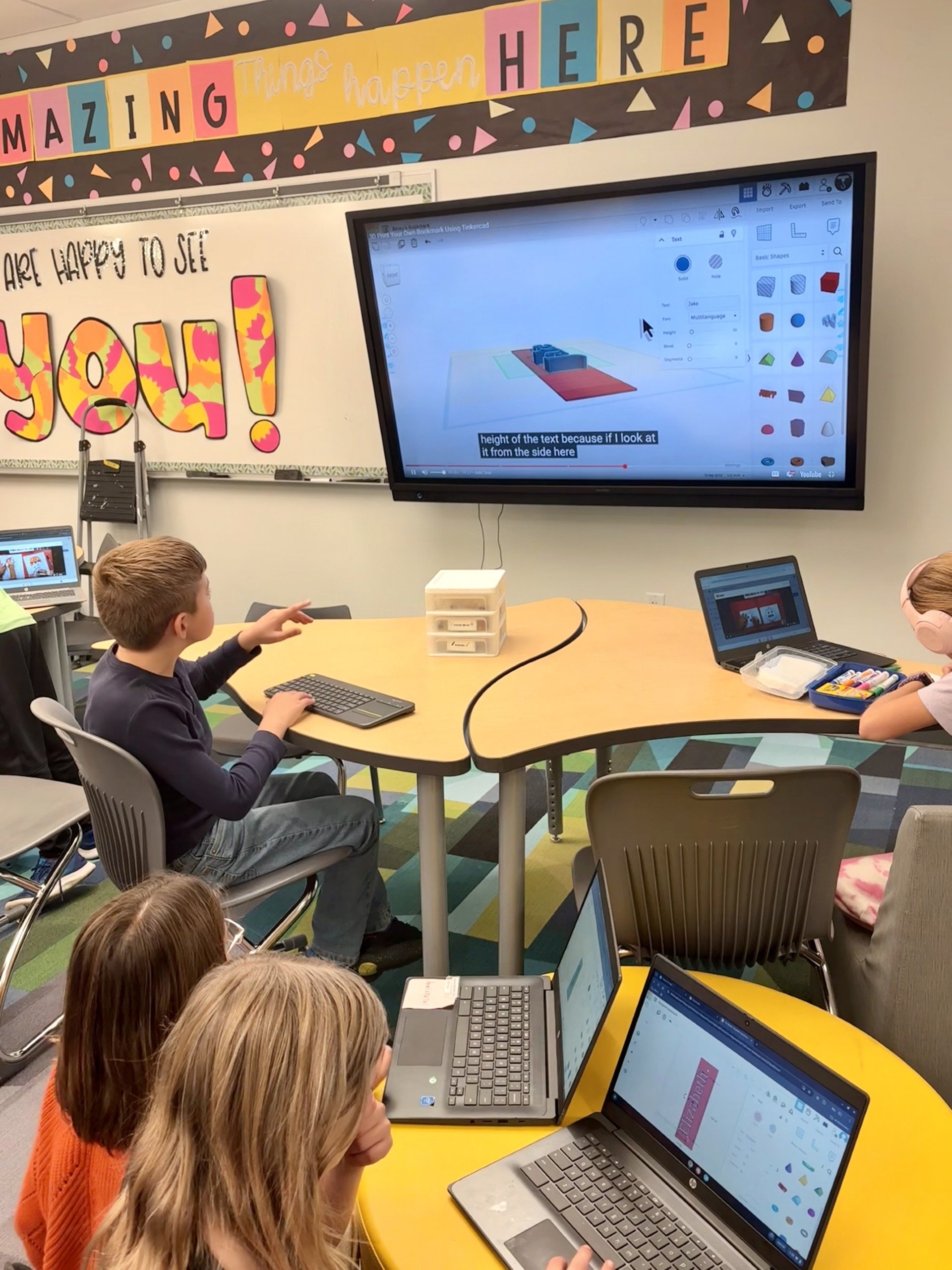 Children working on laptops and a large screen in a classroom, with colorful wall decorations and a banner that says 'AMAZING things happen HERE' and a sign that says 'You are happy to see joy!'
