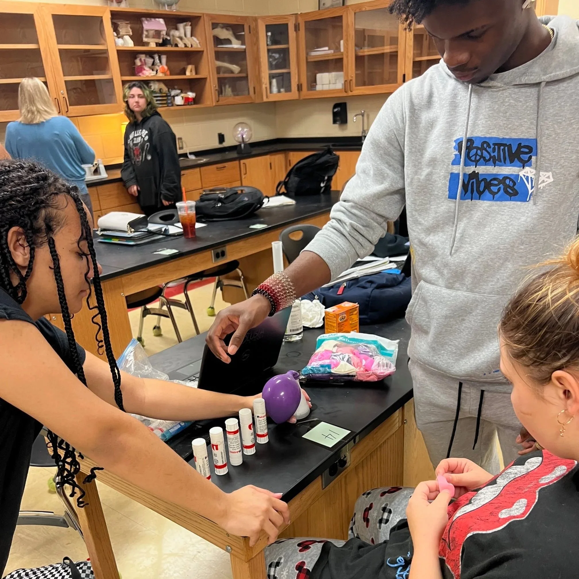 Underserved students gathered around a science classroom table with lab supplies and snacks, one student working on a project while others look on, and additional students in the background near laboratory cabinets.