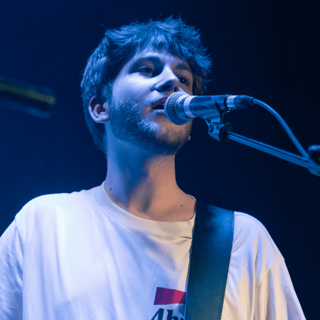 Junger Mann singt in ein Mikrofon auf einer Bühne, blaue Beleuchtung, mit Gitarre. 