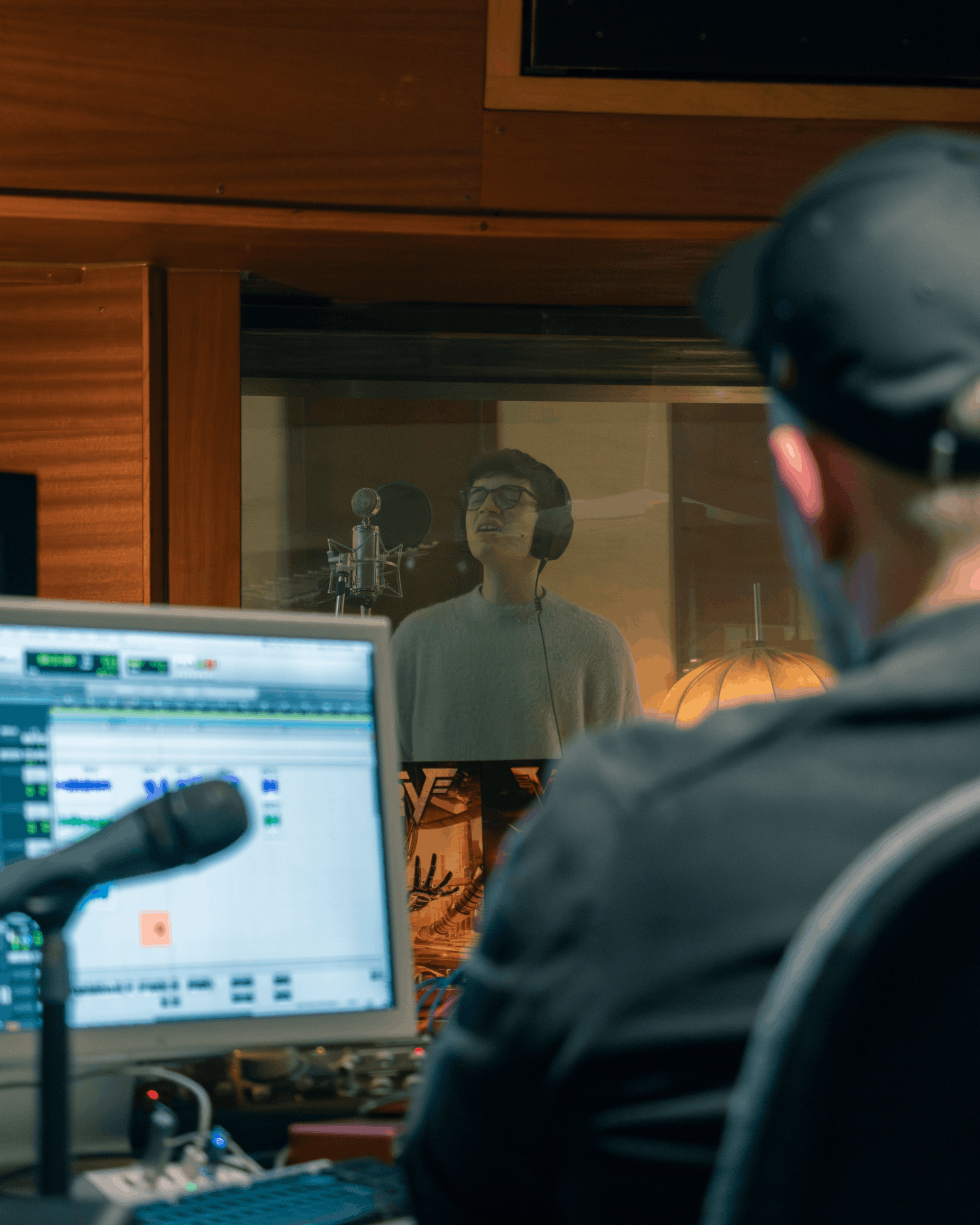 Ein Mann im Tonstudio singt in einem Mikrofon, während ein Toningenieur vor einem Computer arbeitet.