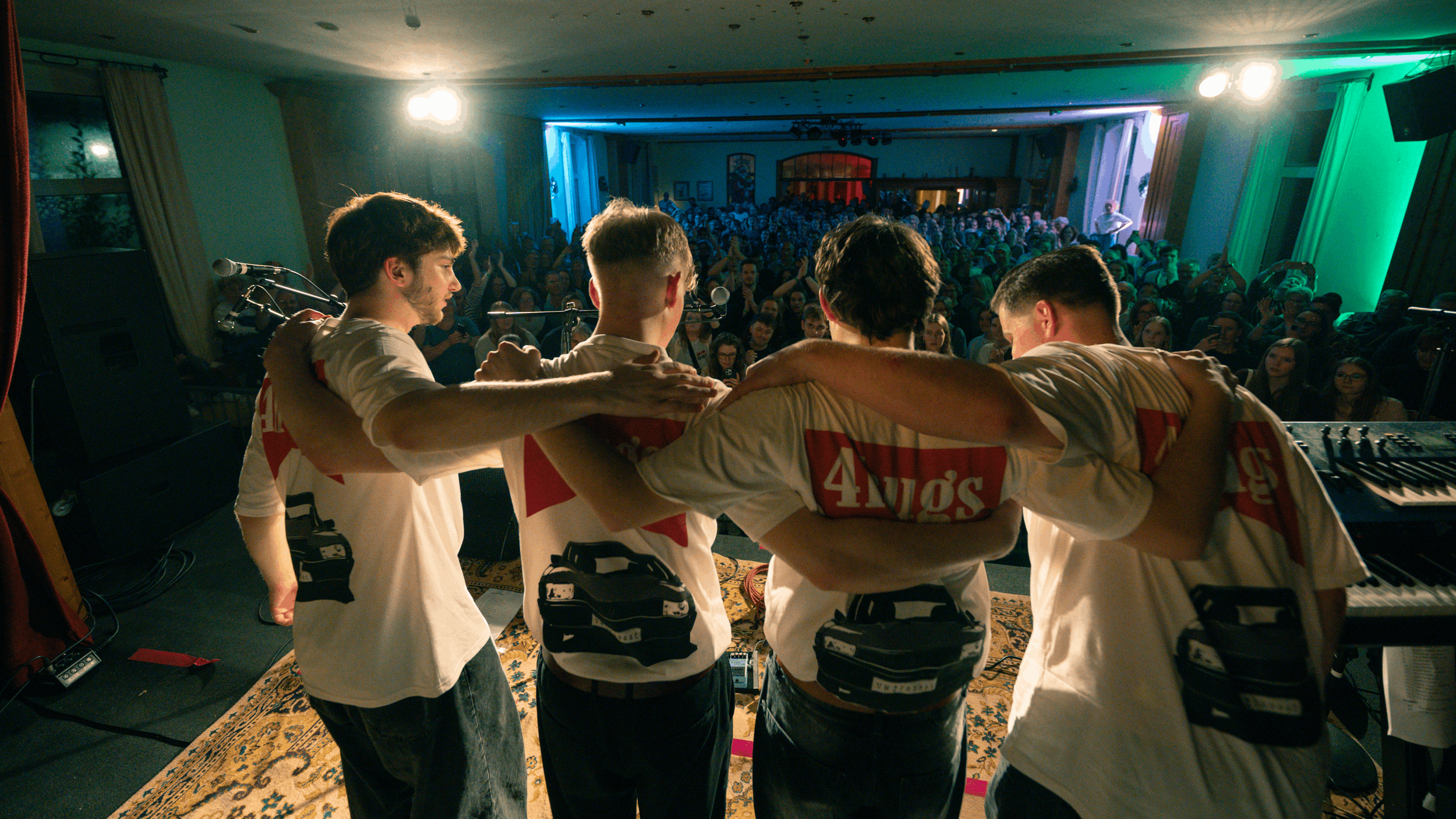 Vier junge Musiker stehen auf der Bühne und umarmen sich, während sie ein Konzert vor einem großen Publikum geben, das im Saal sitzt und zuschaut.