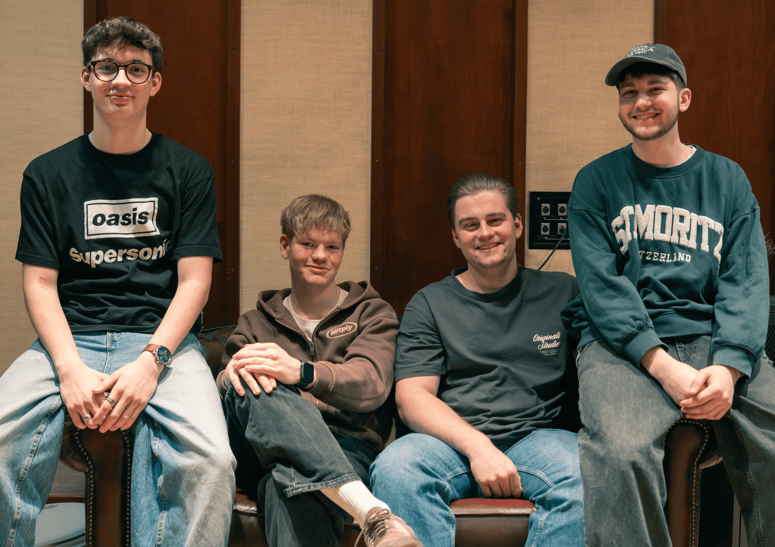 Fünf junge Männer sitzen auf einem roten Sofa in einem Tonstudio, lachen und schauen in die Kamera.