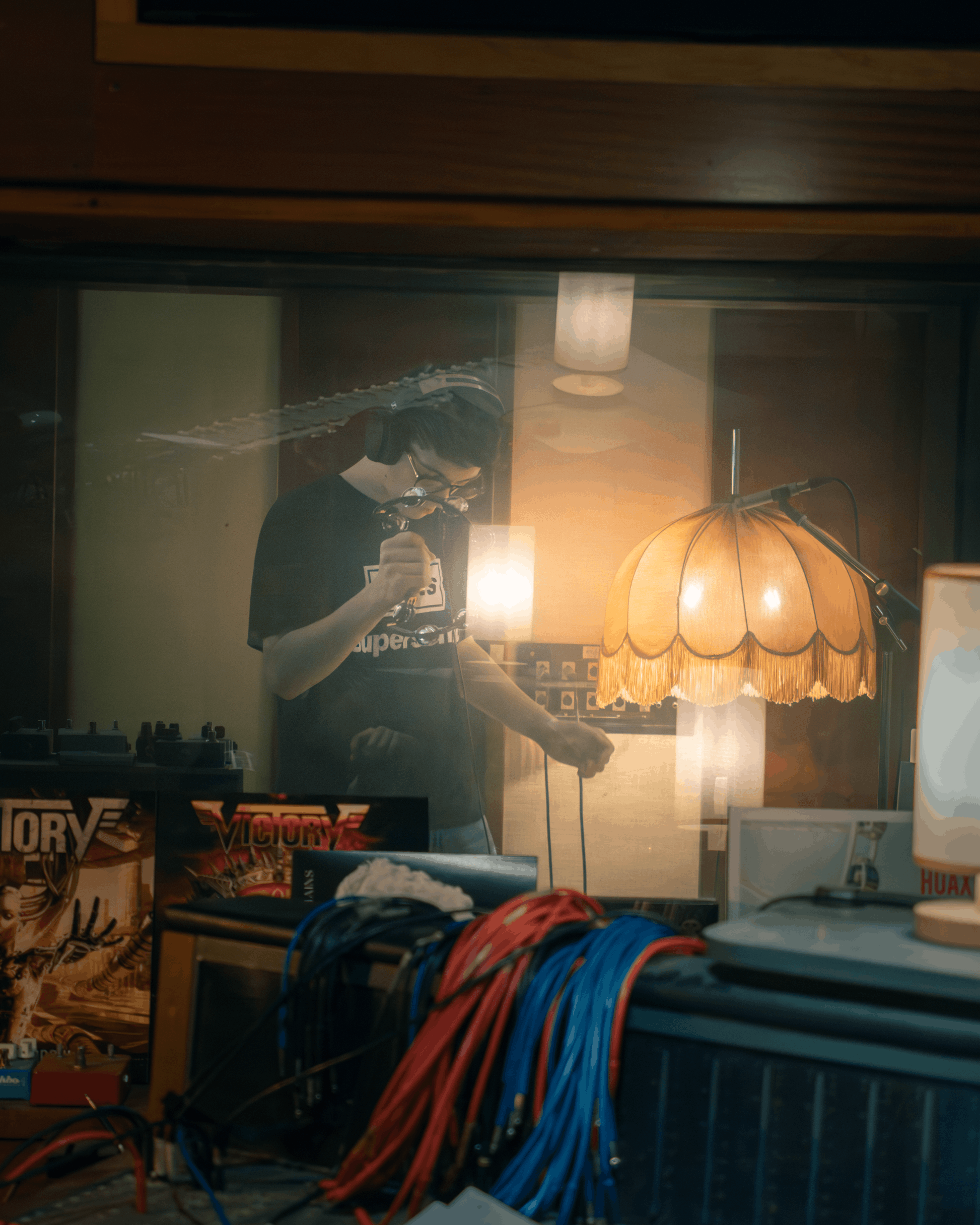Ein junger Mann mit Kopfhörern und Brille am DJ-Pult, hinter einem Fenster, mit einem großen, orangefarbenen Lampenschirm im Hintergrund.