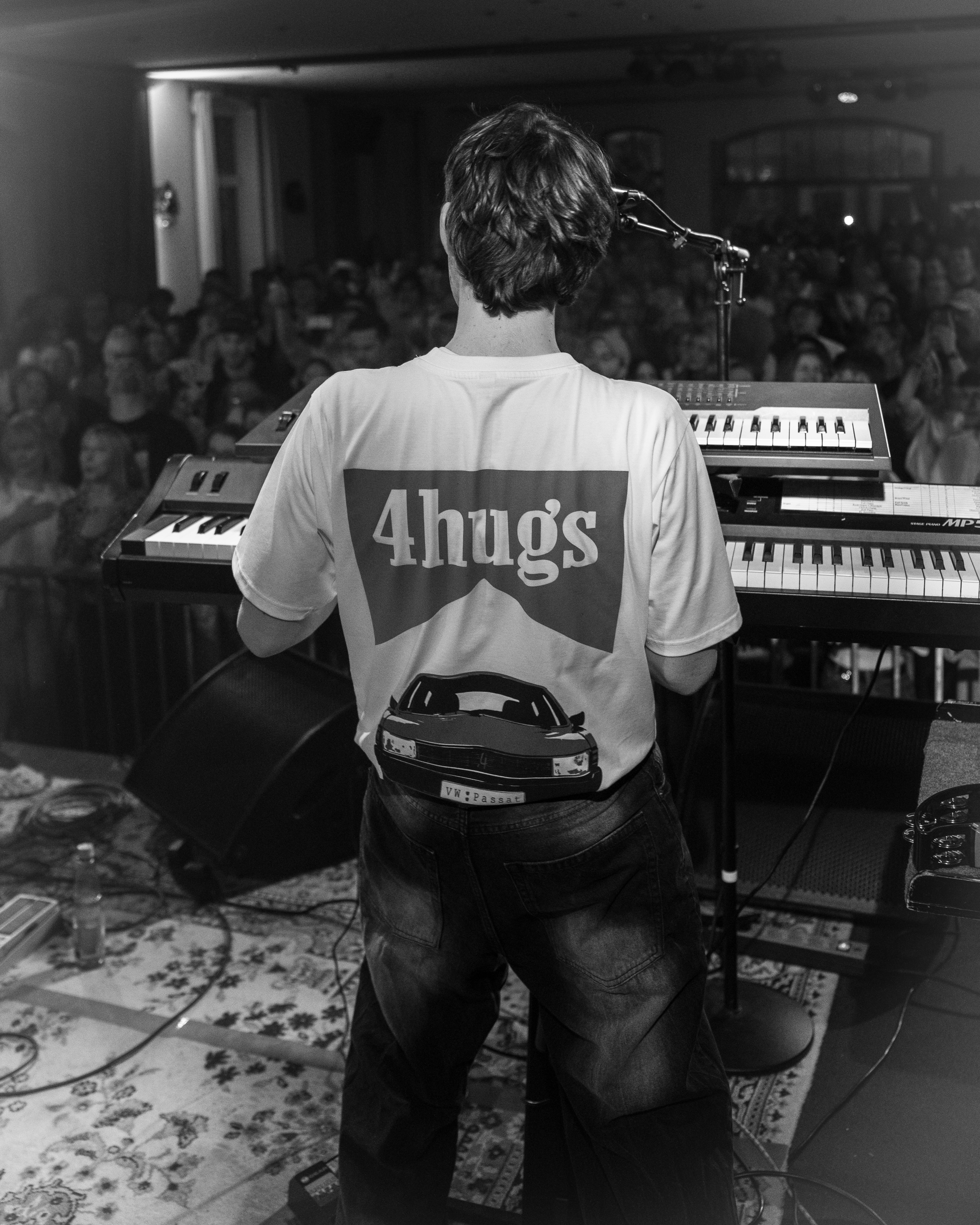 Musiker spielt bei einem Konzert vor einem Publikum, sichtet nur den Rücken, trägt ein T-Shirt mit dem Aufdruck '4hugs' und einem Auto, im Raum voller Zuschauer.