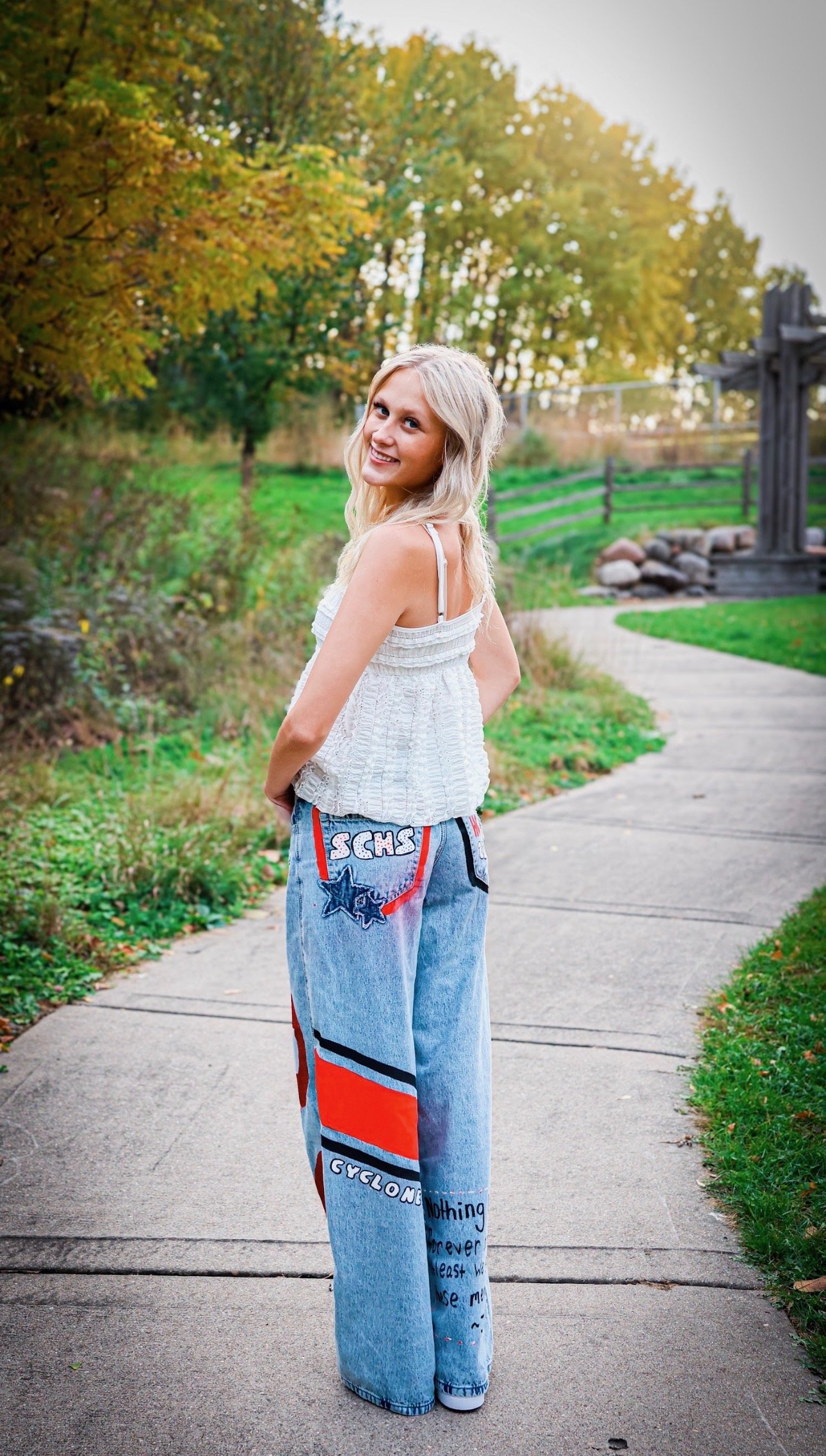 A young woman with long blonde hair smiling and standing on a sidewalk in a park during autumn. She is wearing a white sleeveless top and large loose-fitting jeans with colorful patches and writing. There are trees with yellow and green leaves and a wooden fence in the background.