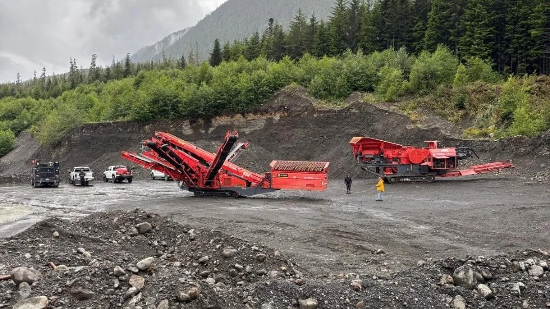 Red aggregate crushing and screening equipment at a gravel site surrounded by forested hills