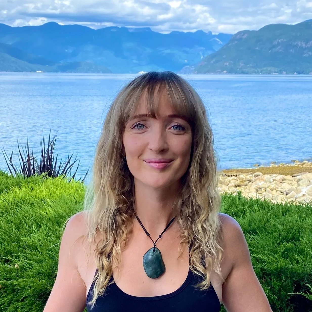 A woman with long curly blonde hair and blue eyes smiling outdoors near a lake, with mountains in the background and green grass in the foreground.