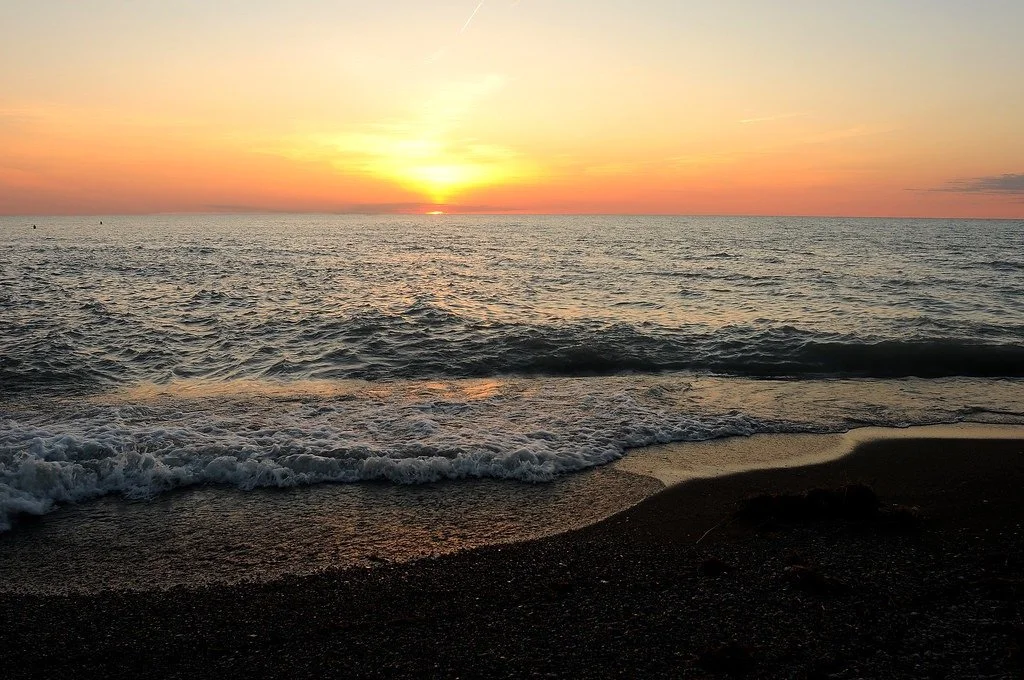 Sunset over the ocean with waves crashing on a dark sandy beach.