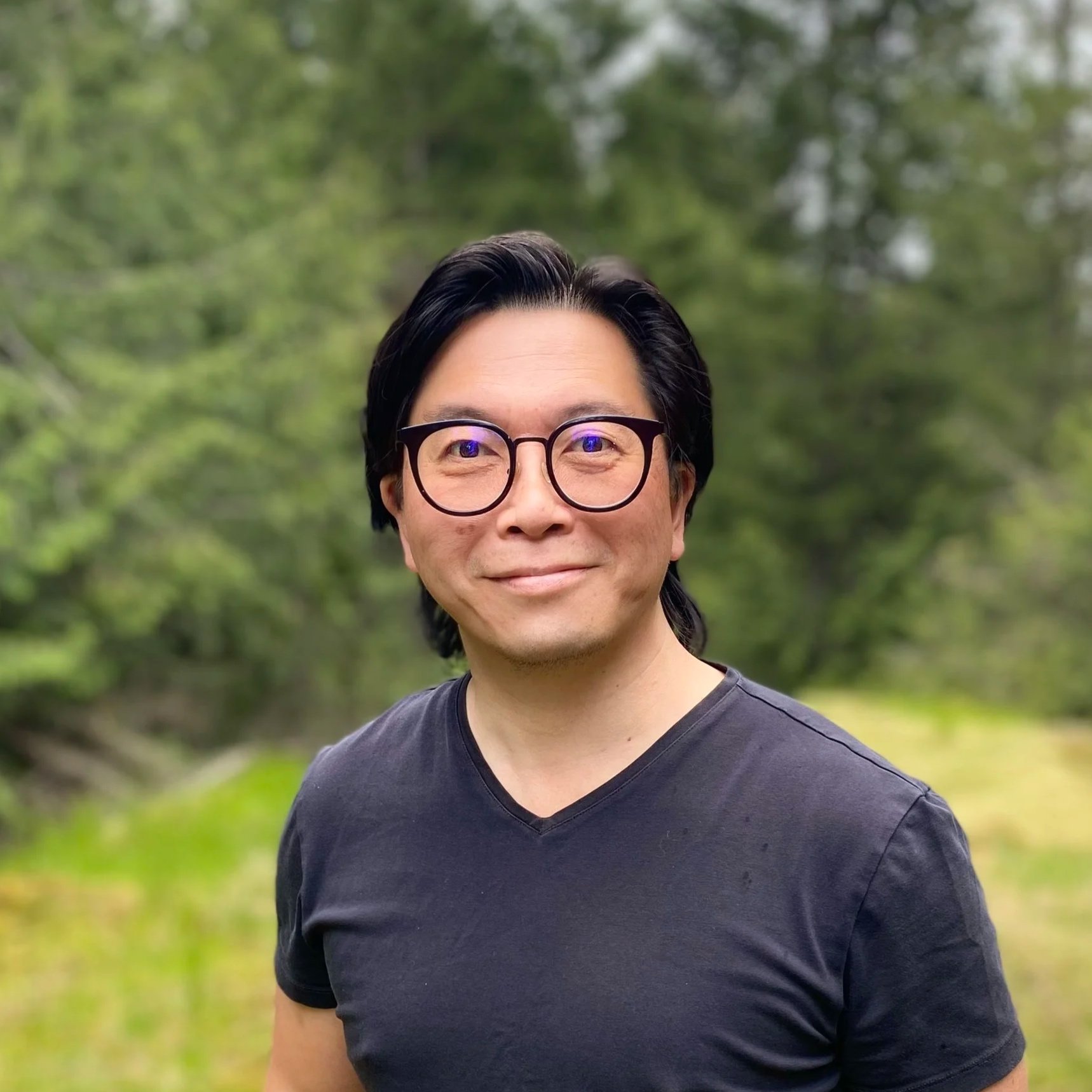 Portrait of a man with black hair, glasses, and a black t-shirt standing outdoors with a blurred green wooded background.