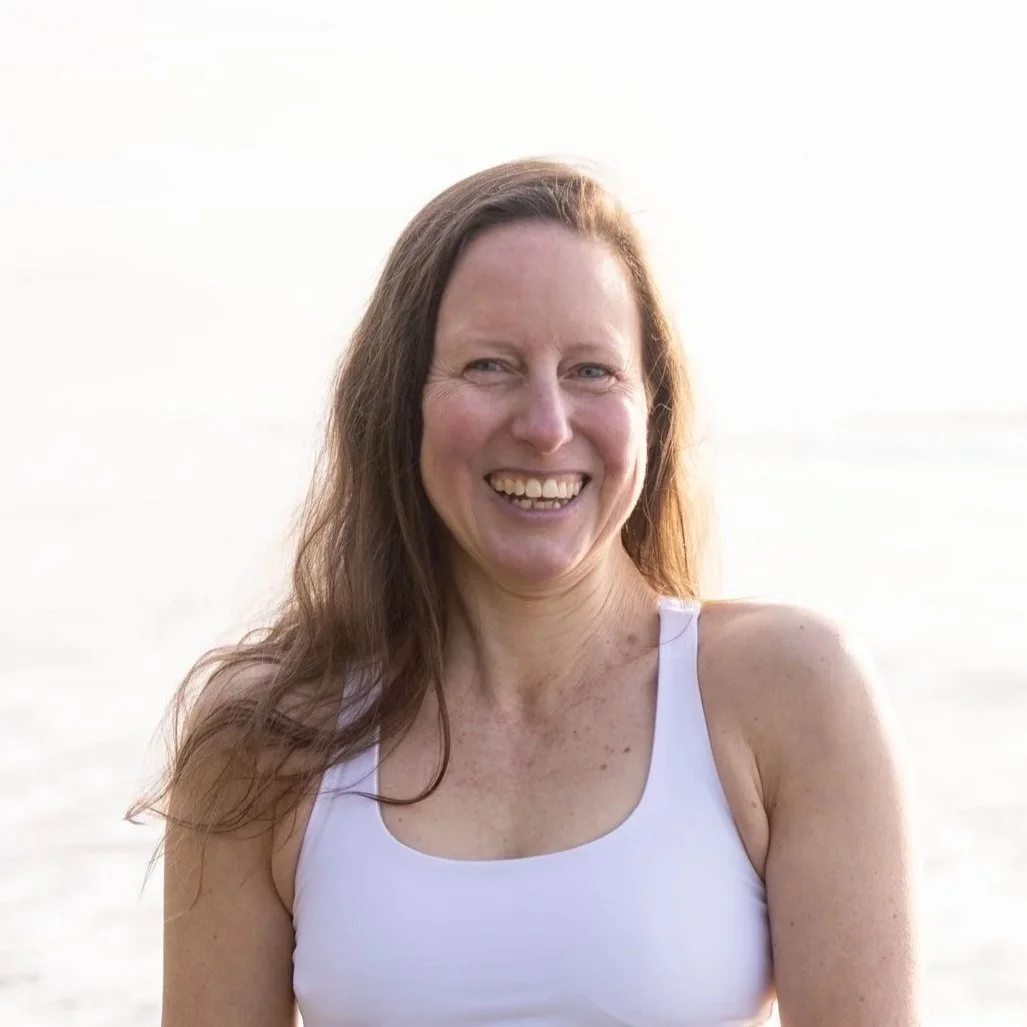 A woman with long brown hair smiling outdoors near water, wearing a white tank top.