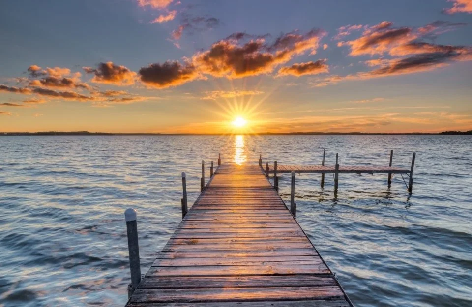 A wooden pier extends into a body of water during sunset, with clouds in the sky and the sun setting on the horizon.