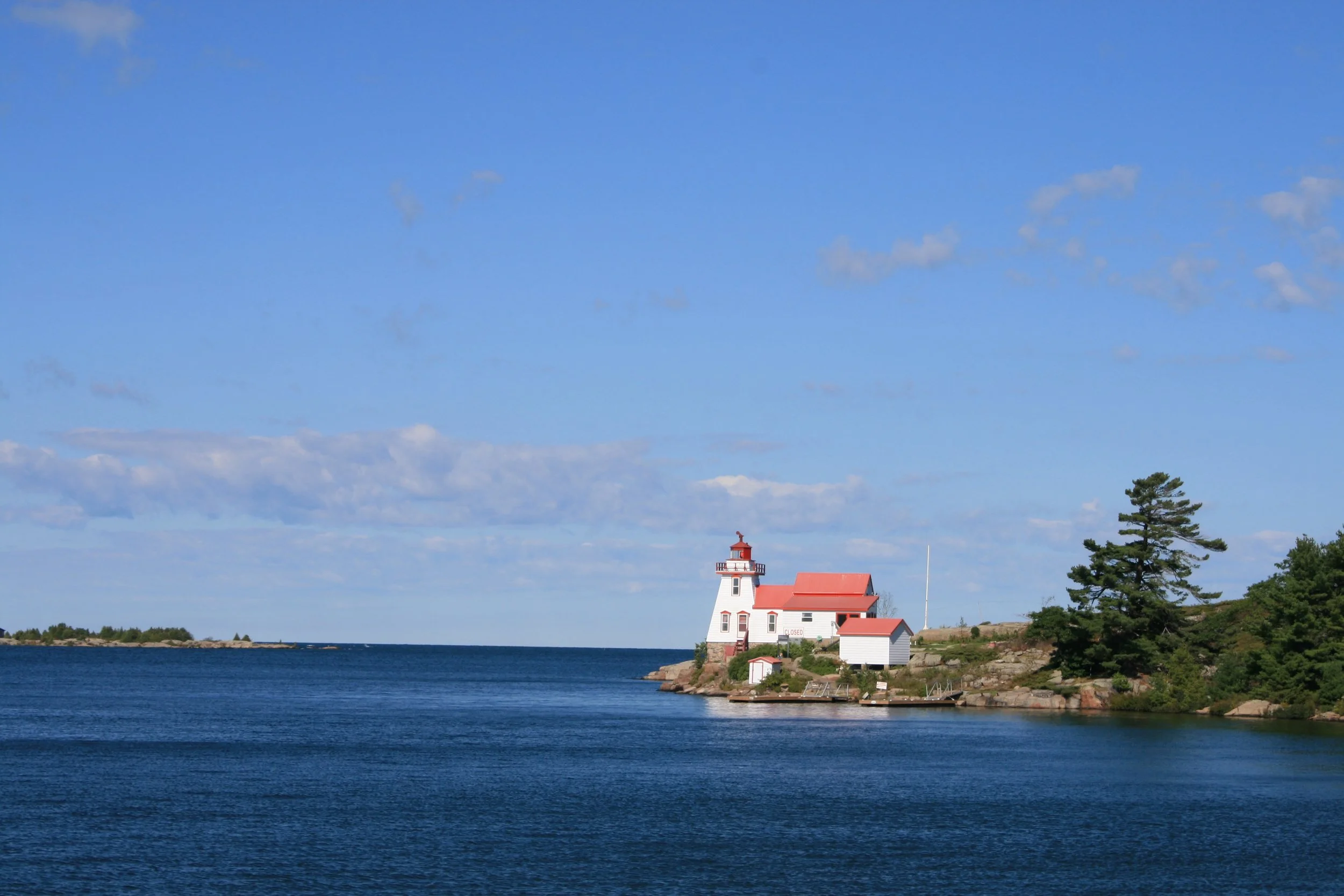A lighthouse on a rocky coastline surrounded by water, with trees and a blue sky with some clouds.