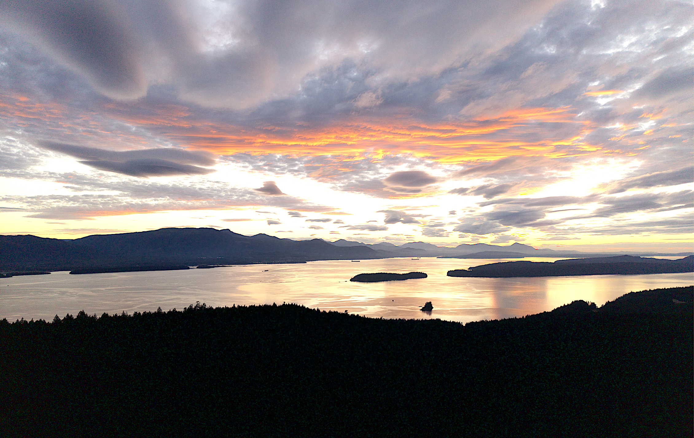 Sunset over a lake with mountains in the background, several boats floating on the calm water.