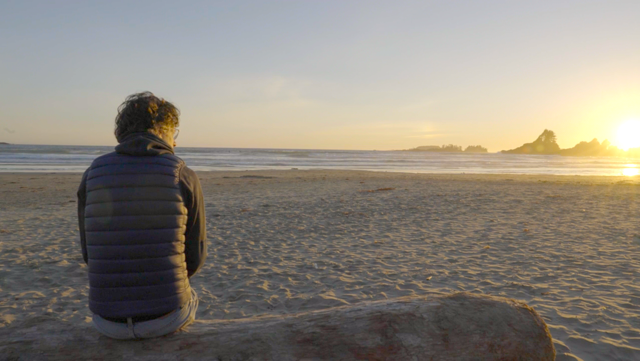 A peaceful beach scene at sunset with wet sand, calm water, trees on a hillside, and an overcast sky.
