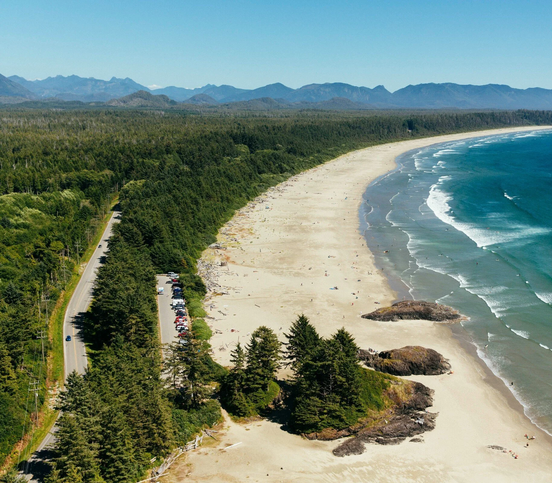 Aerial view of a long sandy beach with rocks, ocean waves, dense green forest, a road with cars, and distant mountains under a clear blue sky.